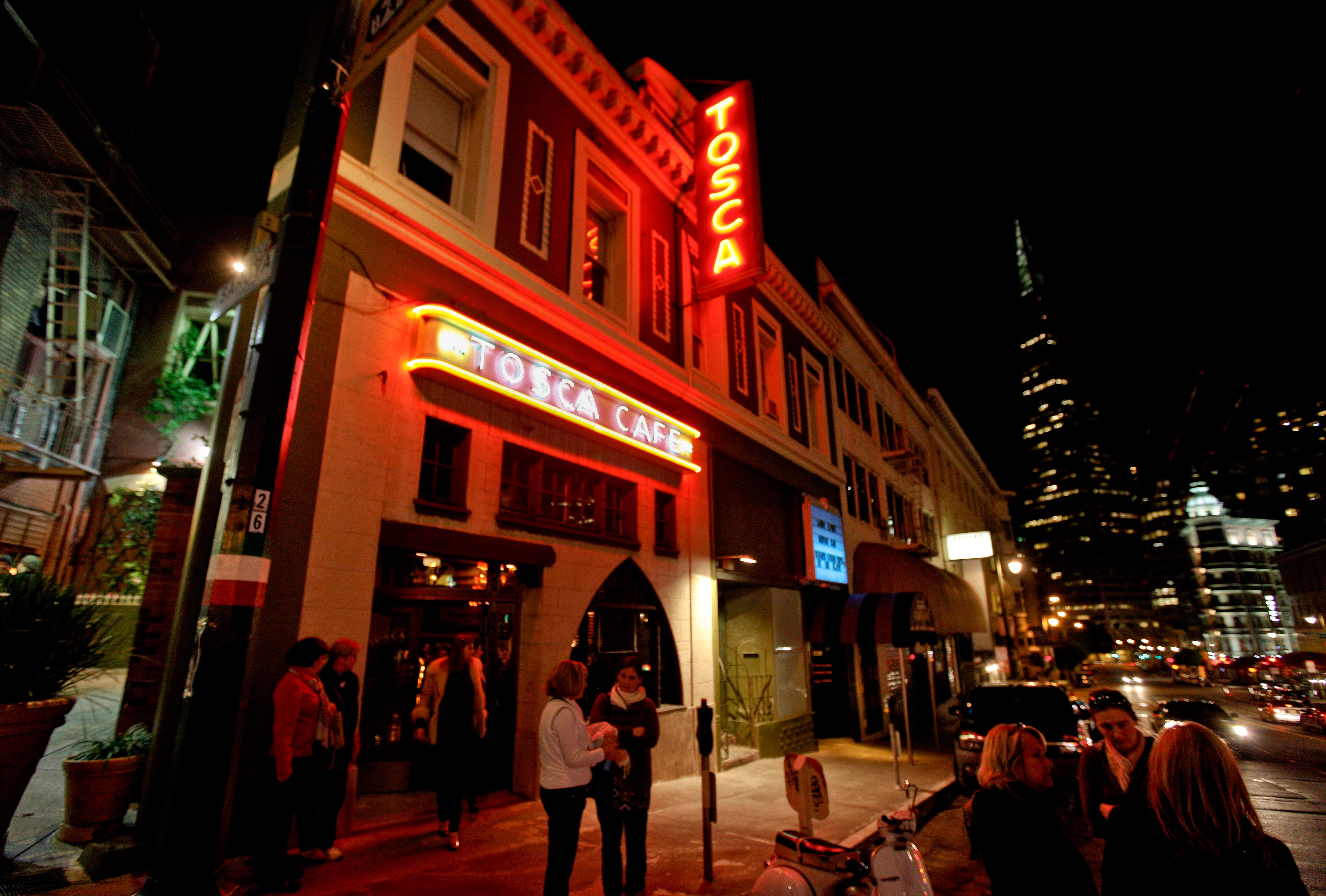 People gather outside a warmly lit Tosca Café at night, with a neon sign and city buildings in the background.