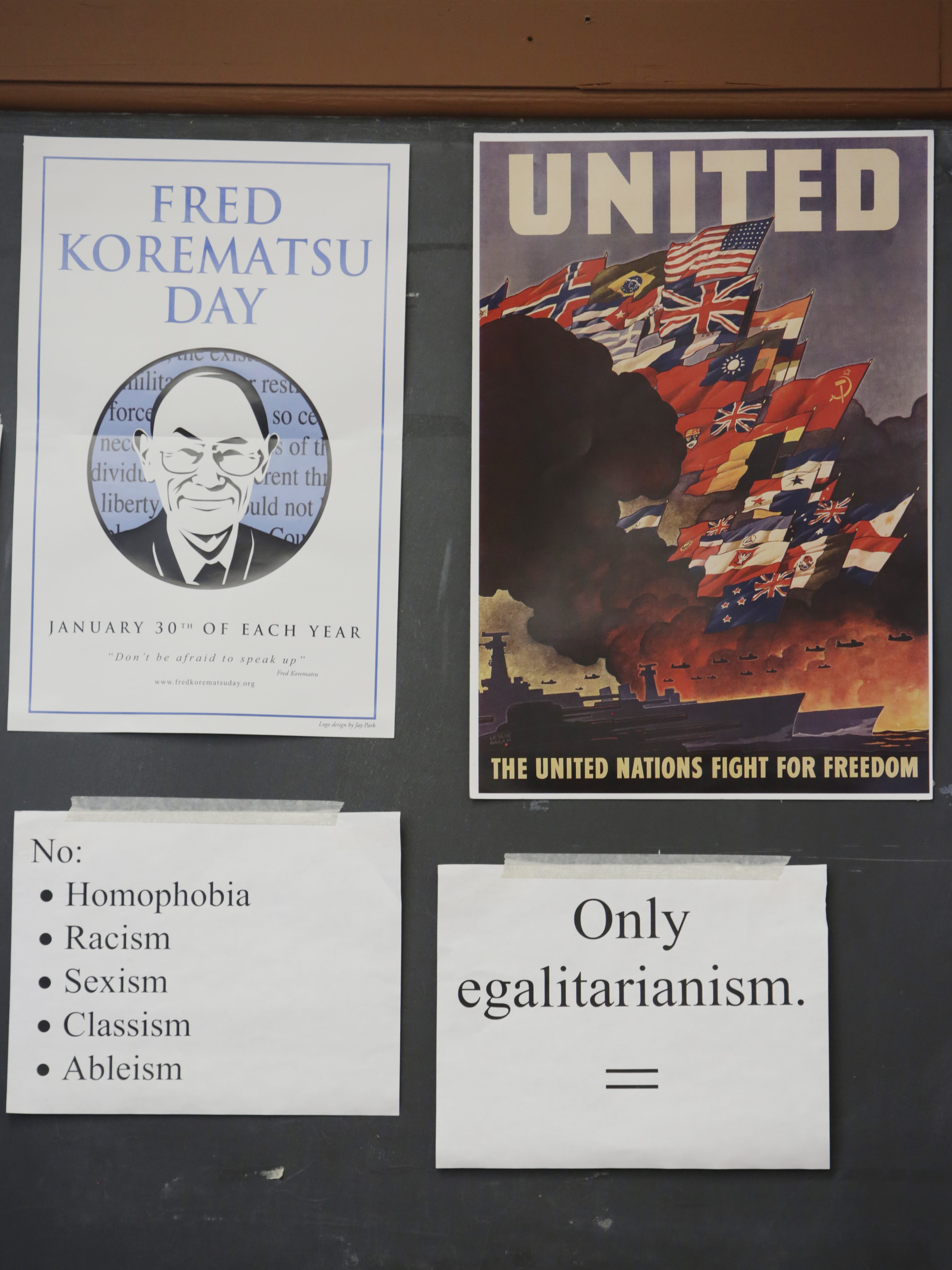 Two posters, one for Fred Korematsu Day, the other showing United Nations flags. Below are signs opposing discrimination and supporting egalitarianism.
