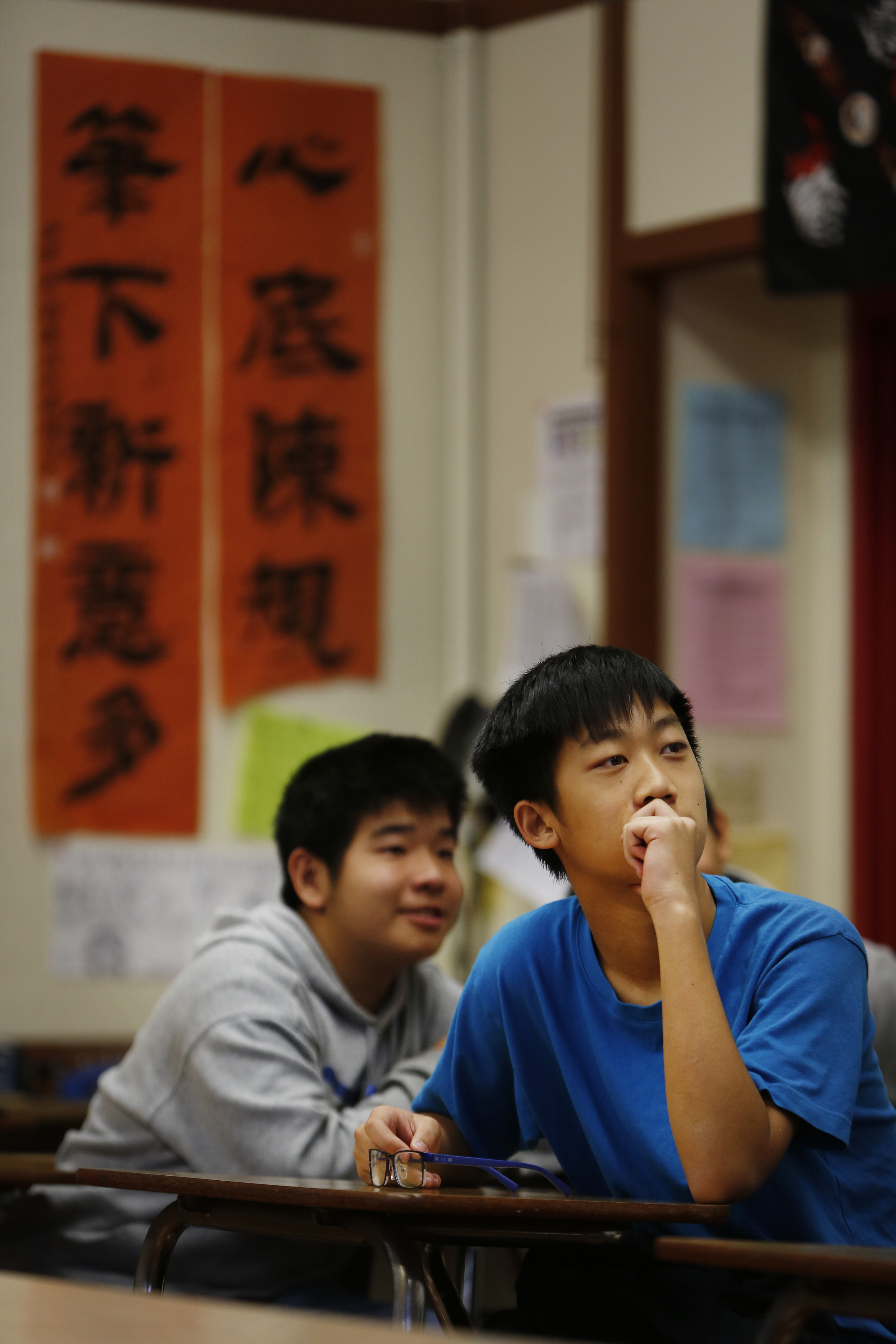 Two students sit attentively in a classroom, one wearing a blue shirt with his hand on his chin, and the other in a gray hoodie. Asian calligraphy hangs on the wall.