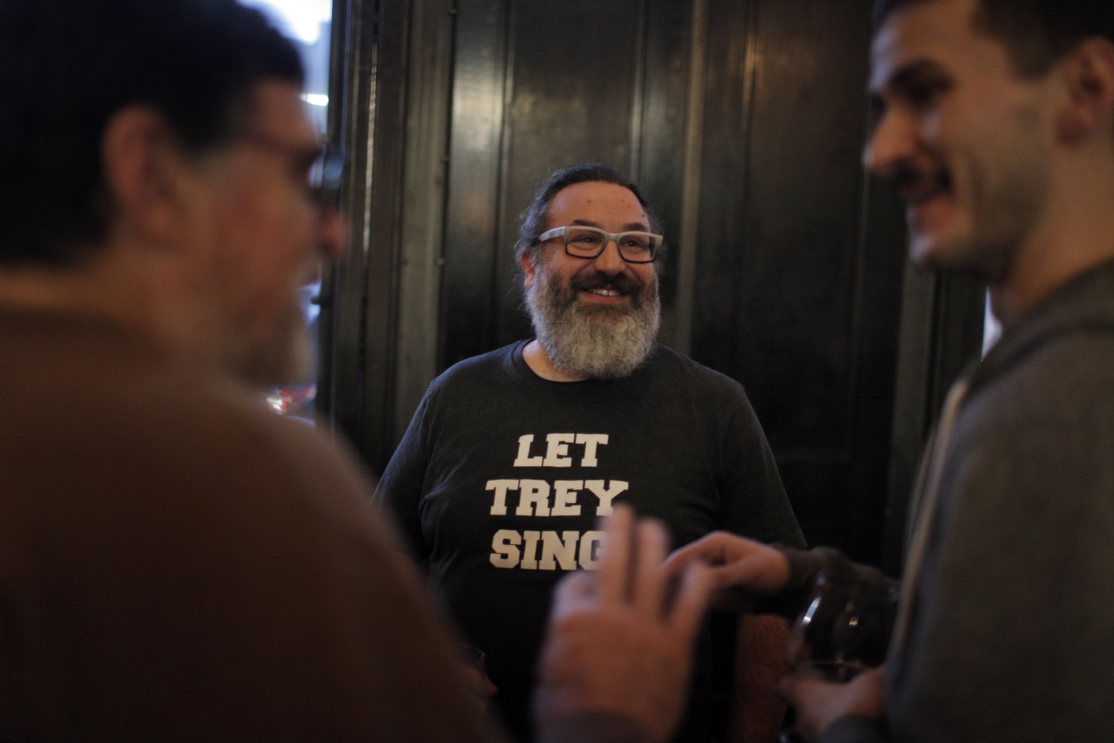 Three men are in conversation; the man in the center has a beard, glasses, and a shirt with "LET TREY SING" printed on it. They appear to be in a dimly lit room.
