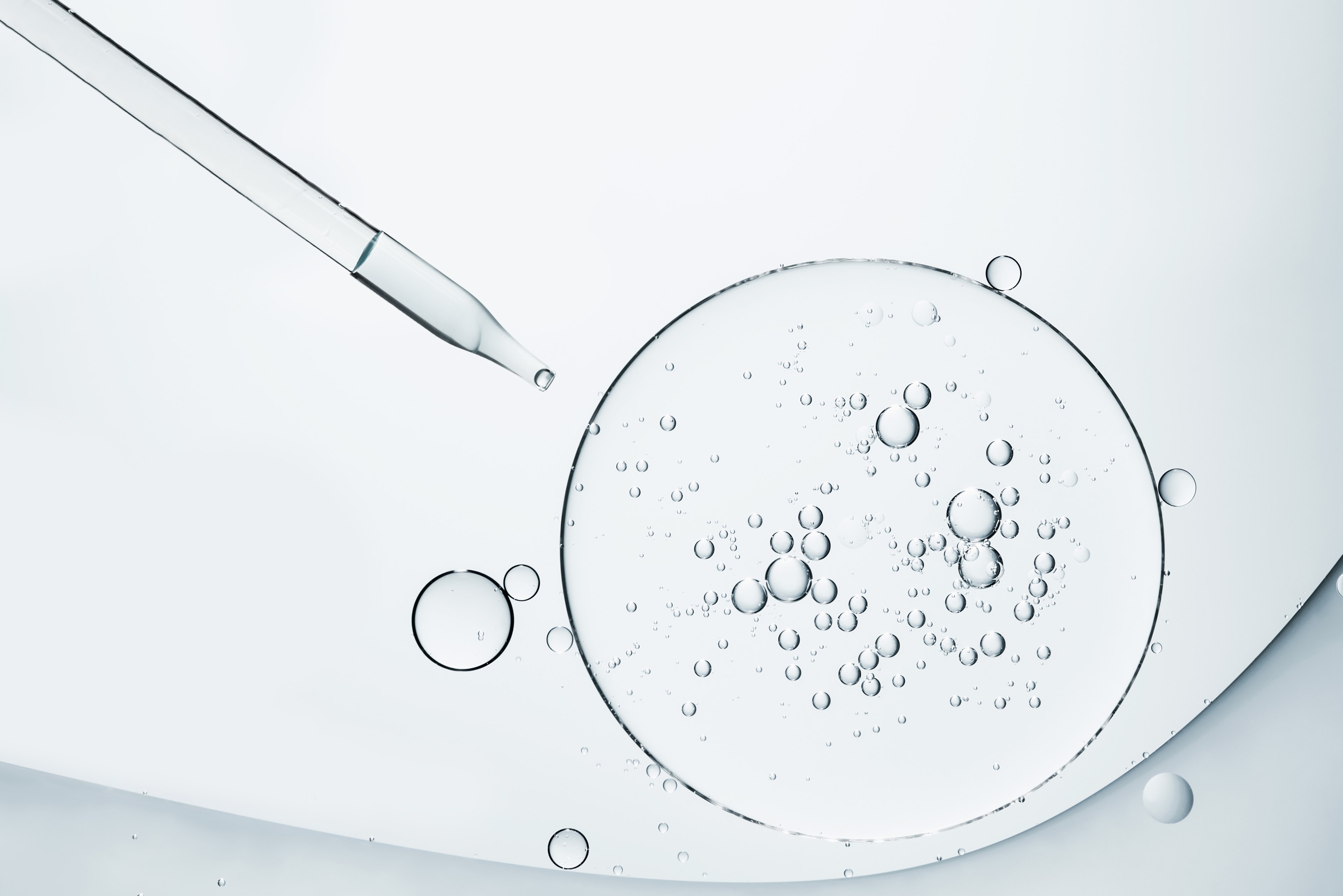 The image shows a clear liquid-filled dropper above large circular surfaces containing bubbles on a smooth, light background.