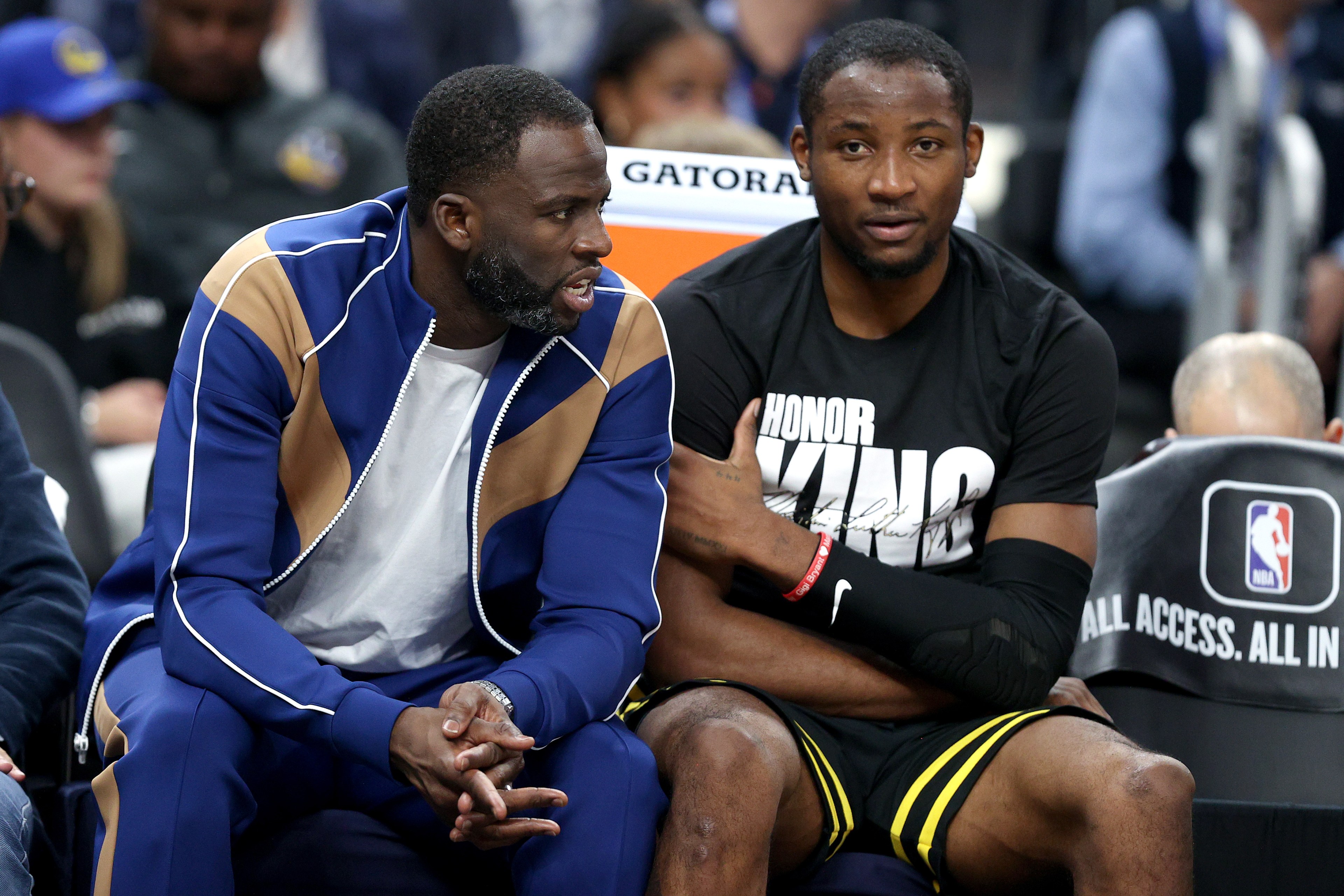Two men sit on a sports bench. One wears a blue and tan tracksuit, talking animatedly. The other in a black &quot;Honor King&quot; shirt listens quietly.