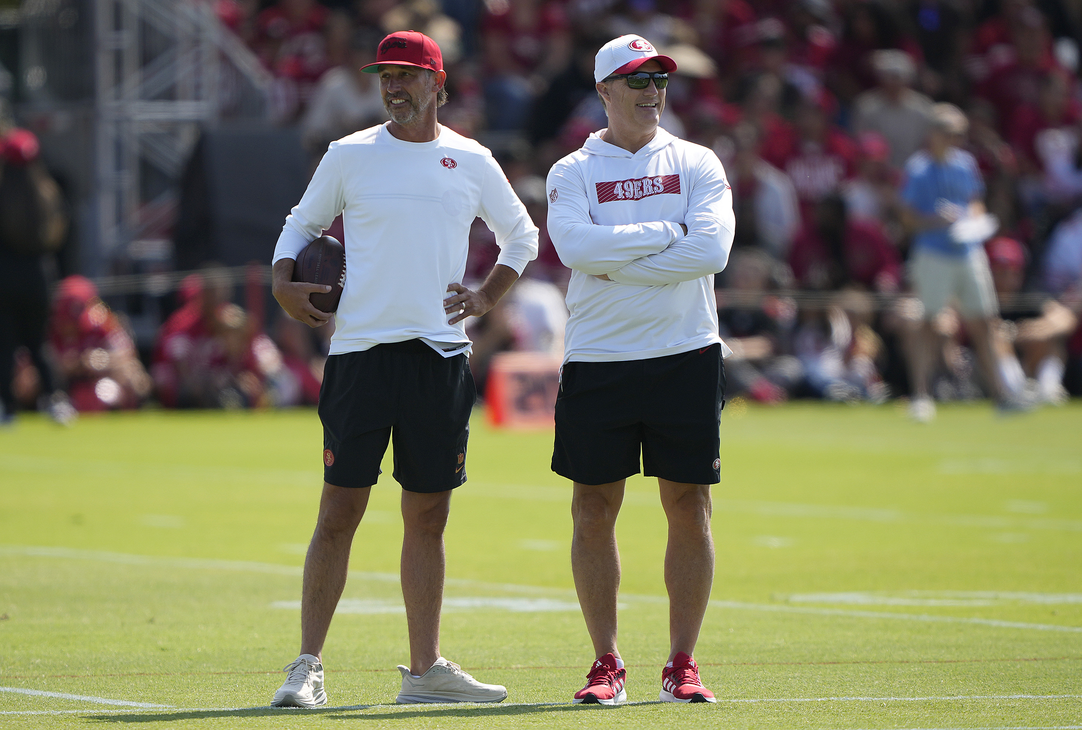Two men in athletic attire stand on a field. One has a red cap and holds a football, while the other wears sunglasses and a white hat. A crowd is in the background.