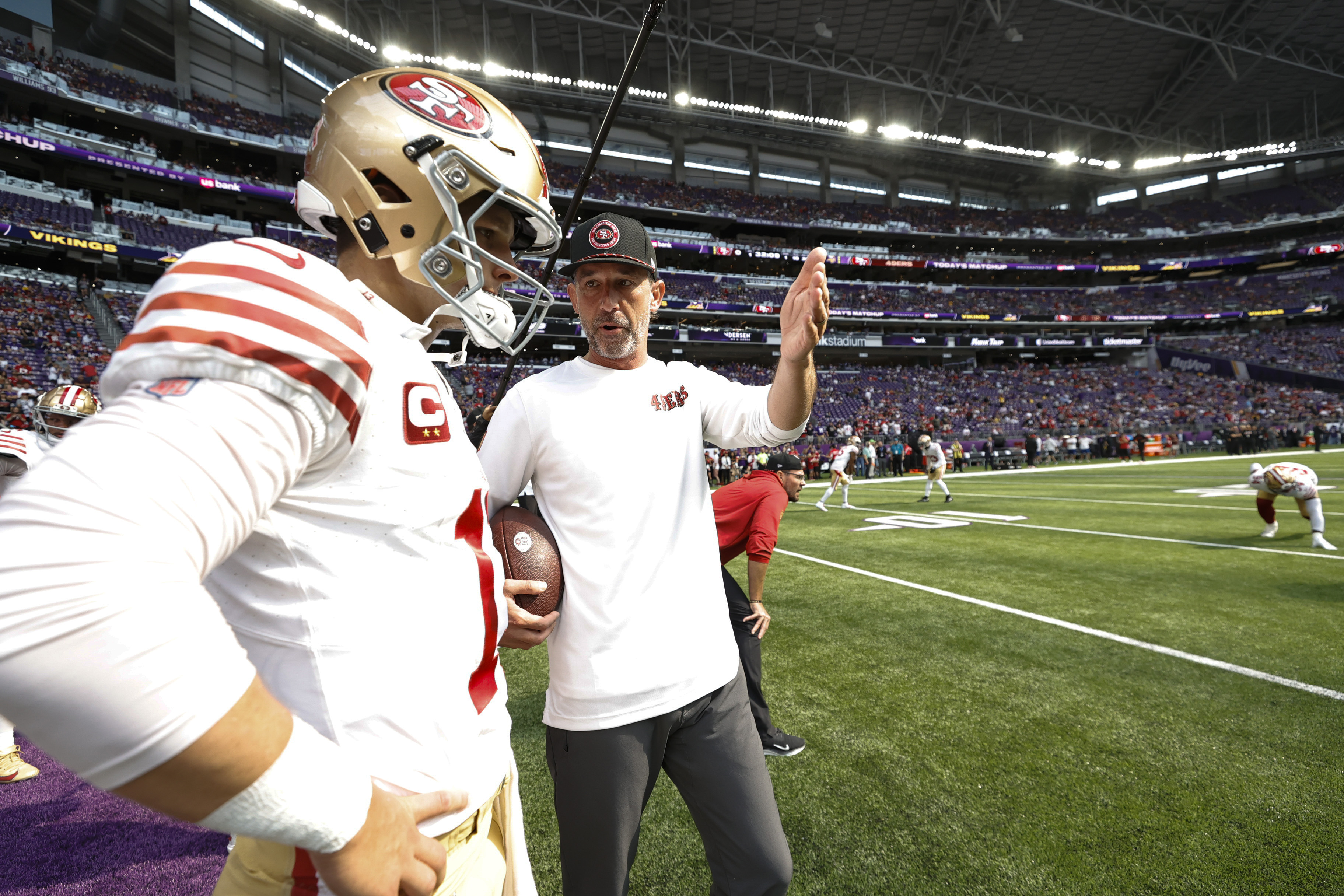 A football coach discusses strategy with a player on the field. The player wears a helmet and jersey with a &quot;C&quot; patch, surrounded by a large stadium crowd.