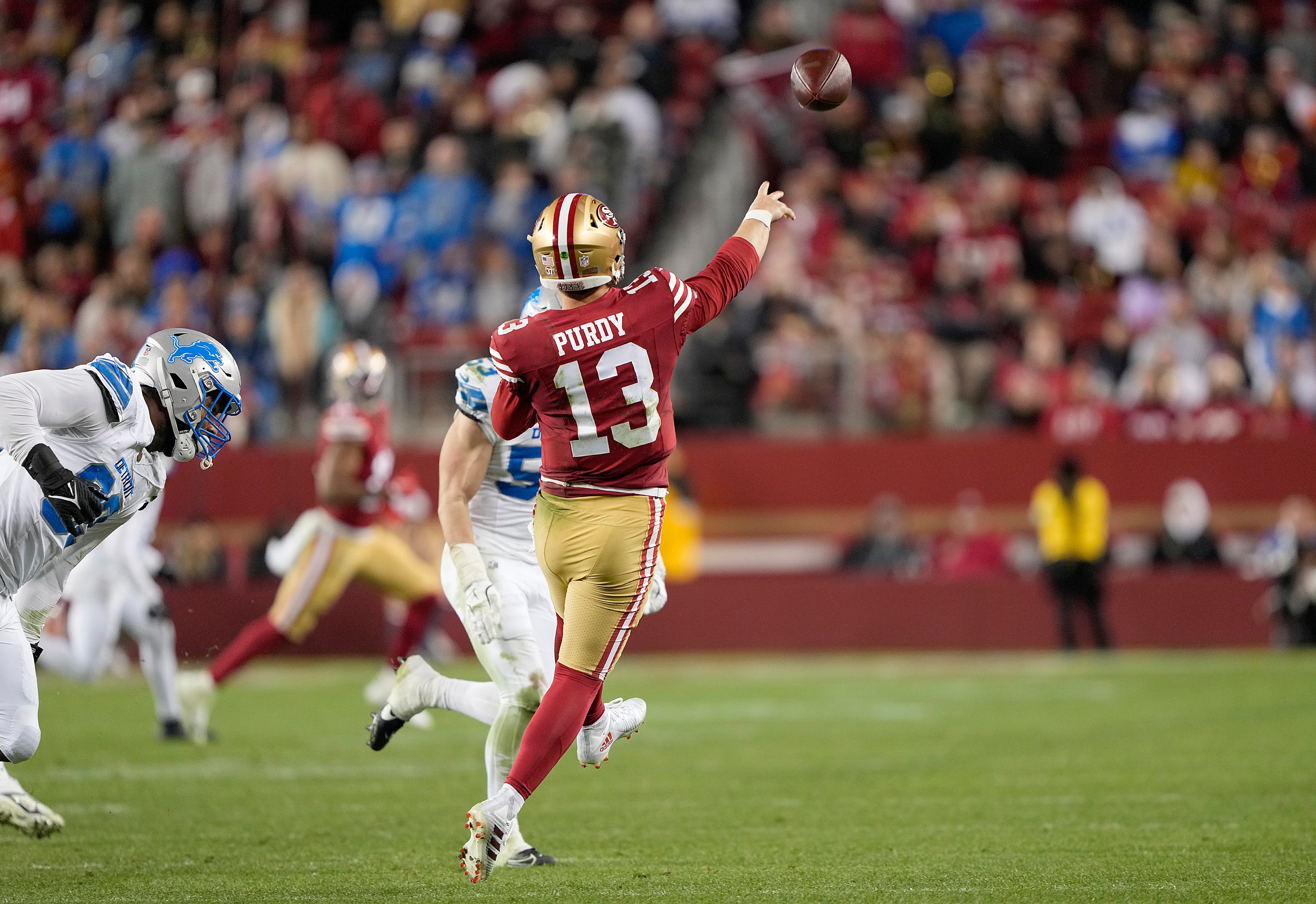 A football player in a red uniform throws a pass while evading a player in a white uniform. The crowd in the background is blurred, focusing on the action.