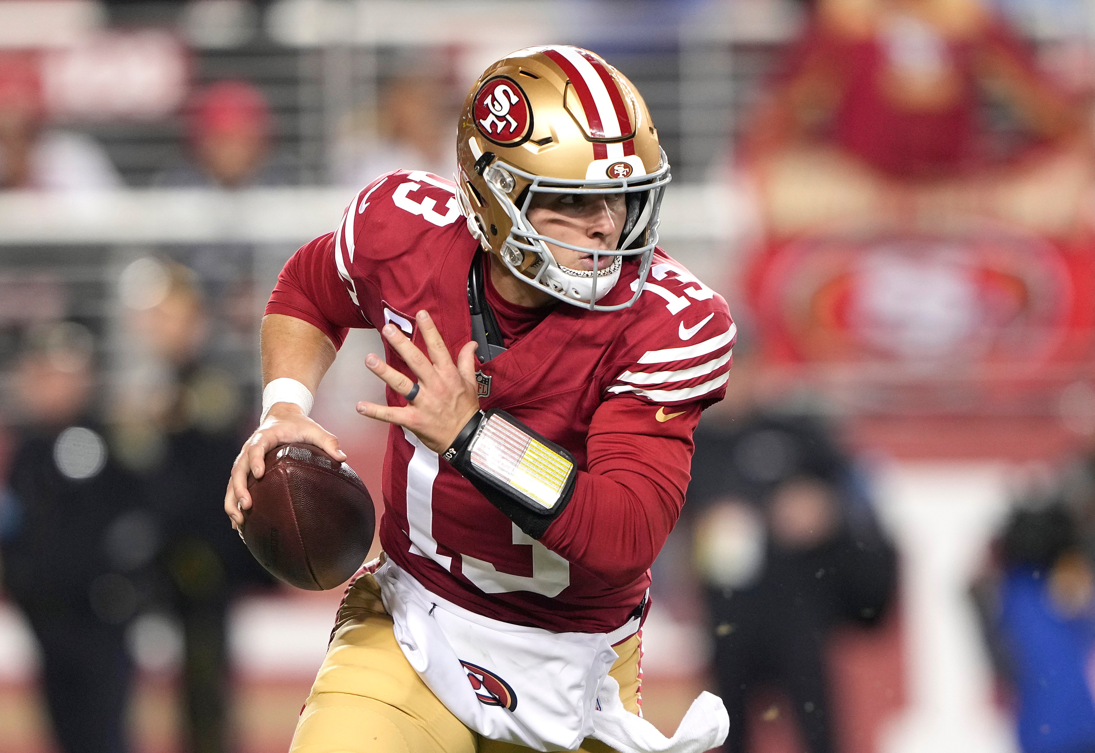 A football player in a red jersey and gold pants runs with the ball, focused and poised. He's wearing a helmet with the number 13 and a team's logo.