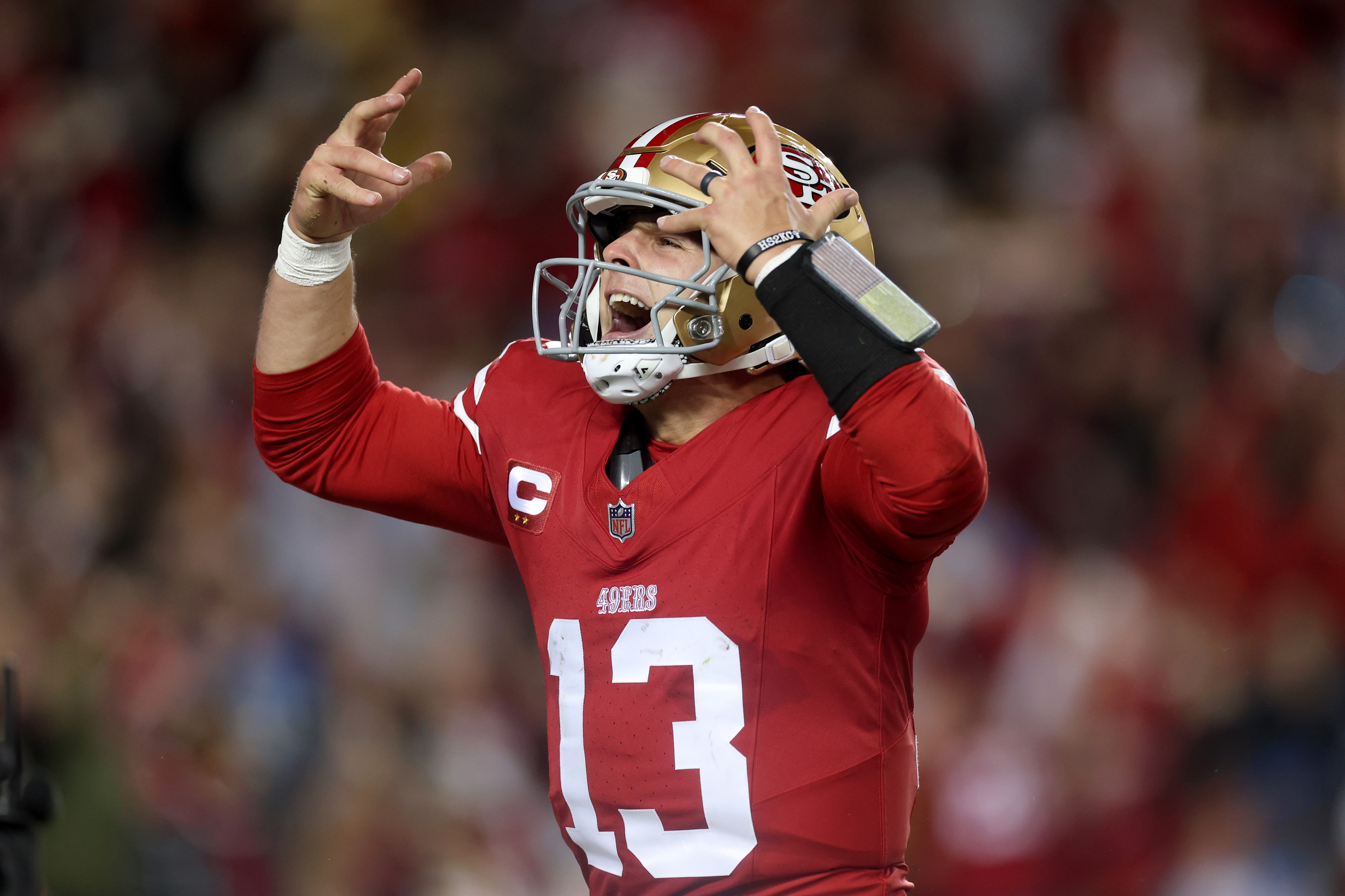 A football player in a red uniform, number 13, is passionately gesturing with his arms raised. He wears a golden helmet and shouts with an intense expression.