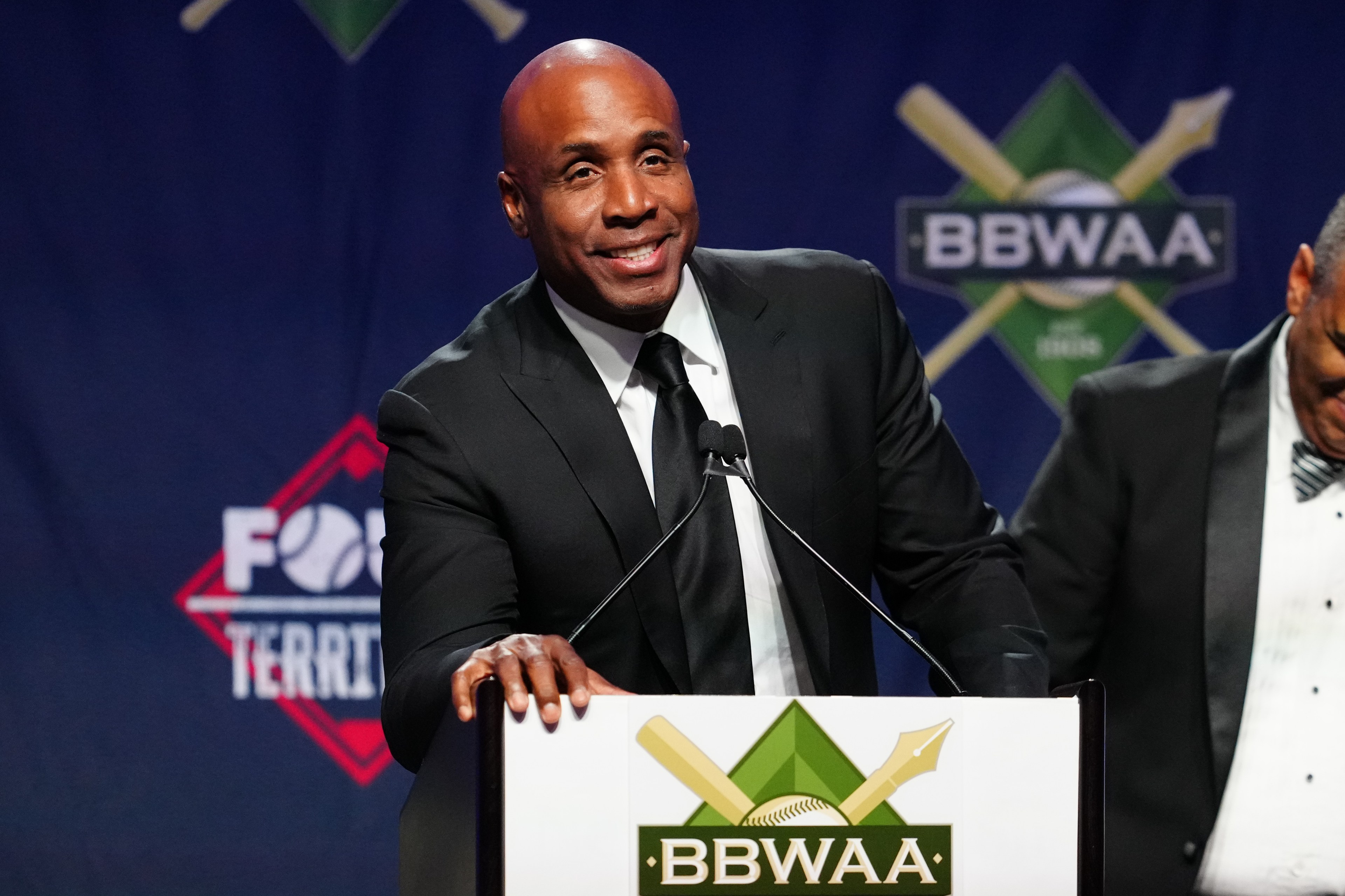 A man in a suit speaks at a podium with a &quot;BBWAA&quot; logo, smiling. A second person stands slightly behind, partially visible, with a blue backdrop.