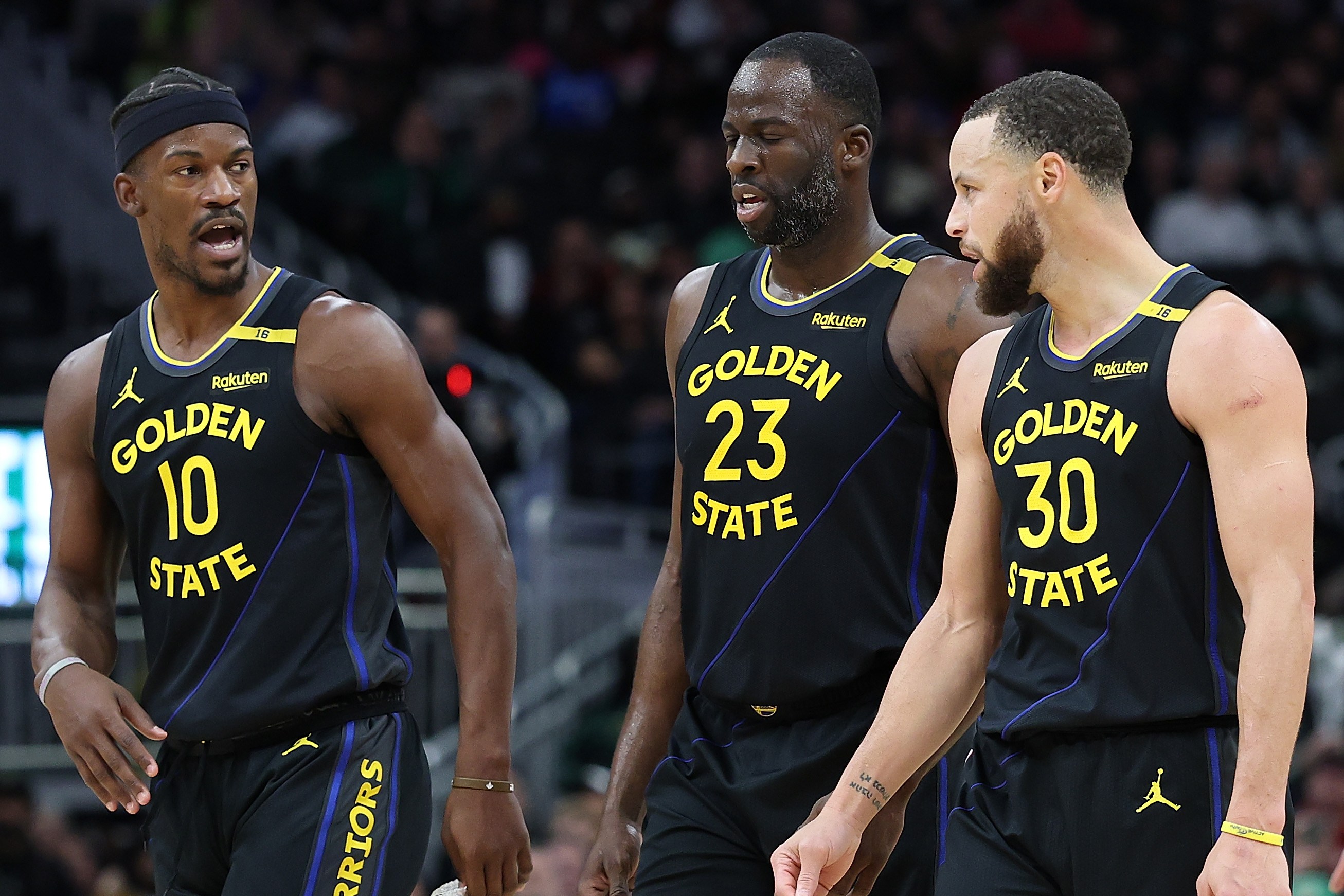 Three basketball players in black "Golden State" uniforms converse on the court, with one gesturing animatedly while walking.