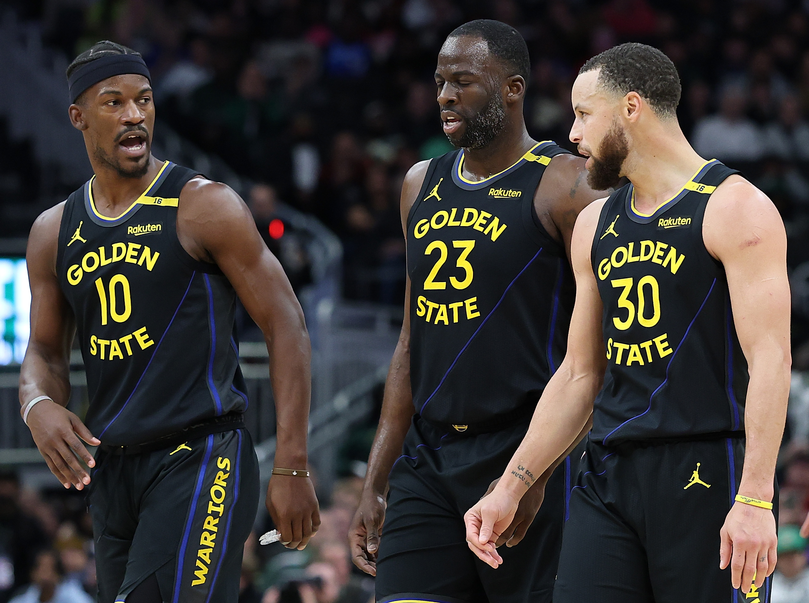 Three basketball players in black "Golden State" uniforms converse on the court, with one gesturing animatedly while walking.
