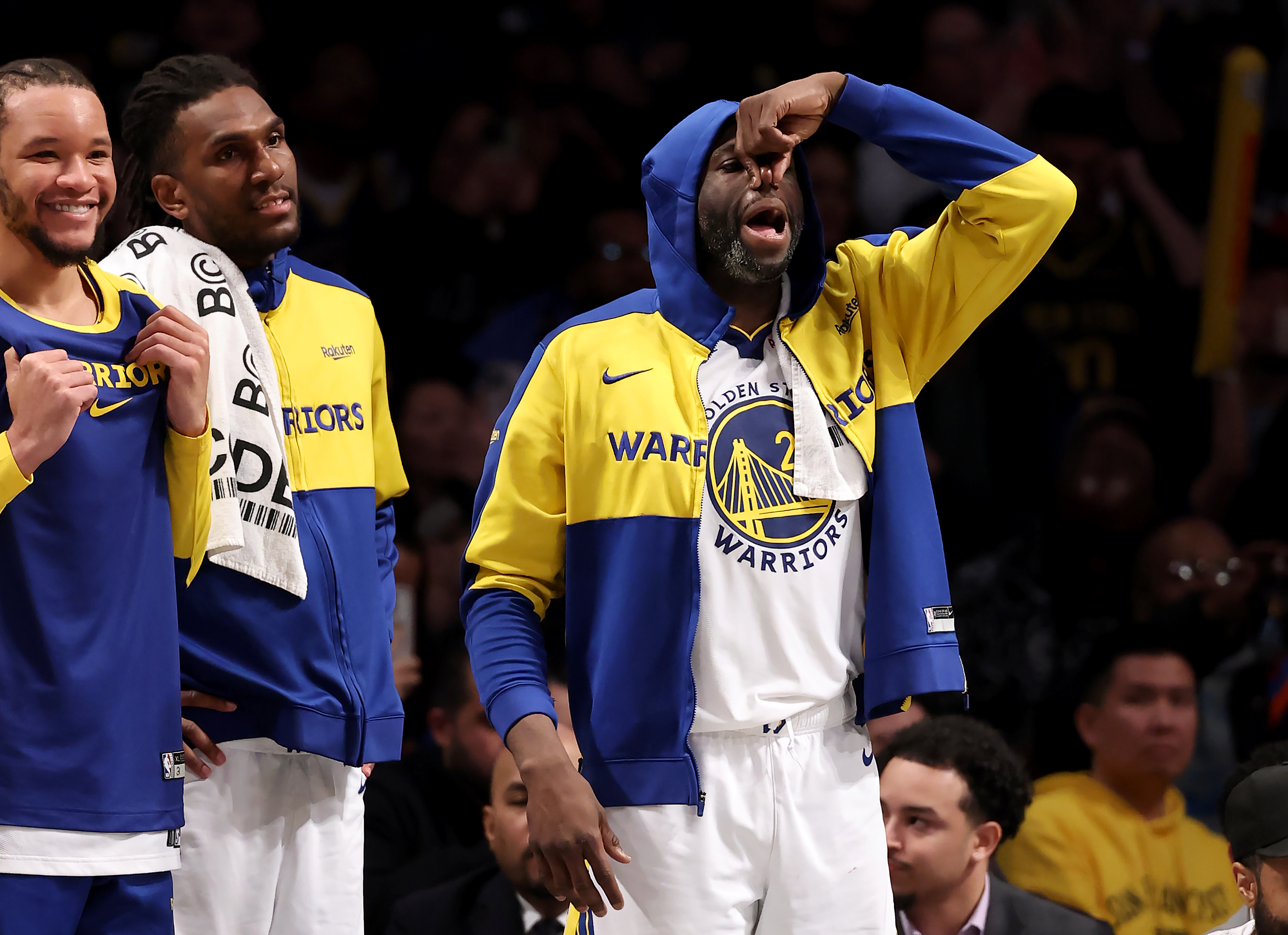 Three basketball players in Warriors gear are on the sideline. One is pinching his nose in a humorous gesture, while the others are smiling and watching.