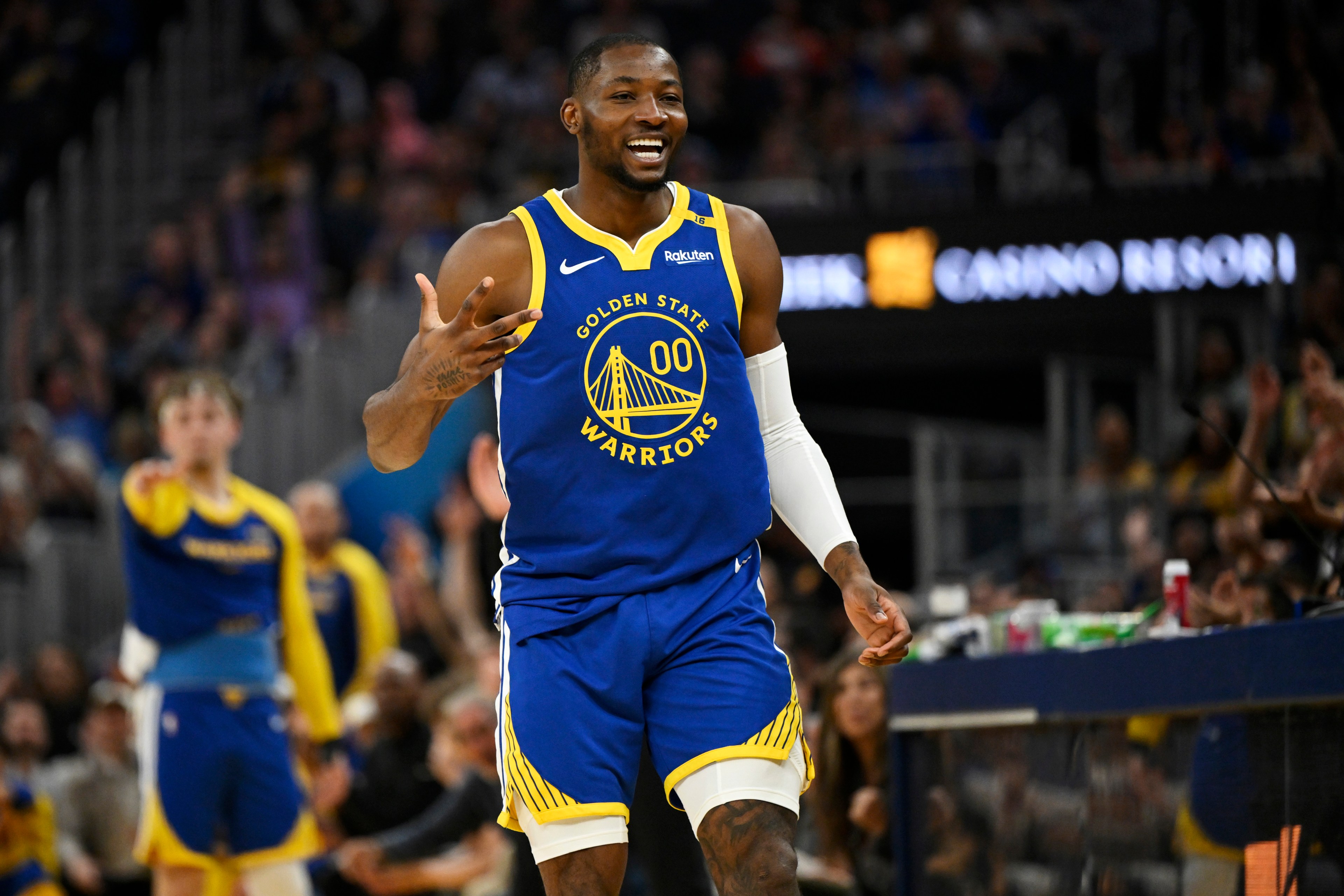 A basketball player in a Golden State Warriors jersey is on the court, smiling and gesturing. The background shows a blurred crowd and part of the court.
