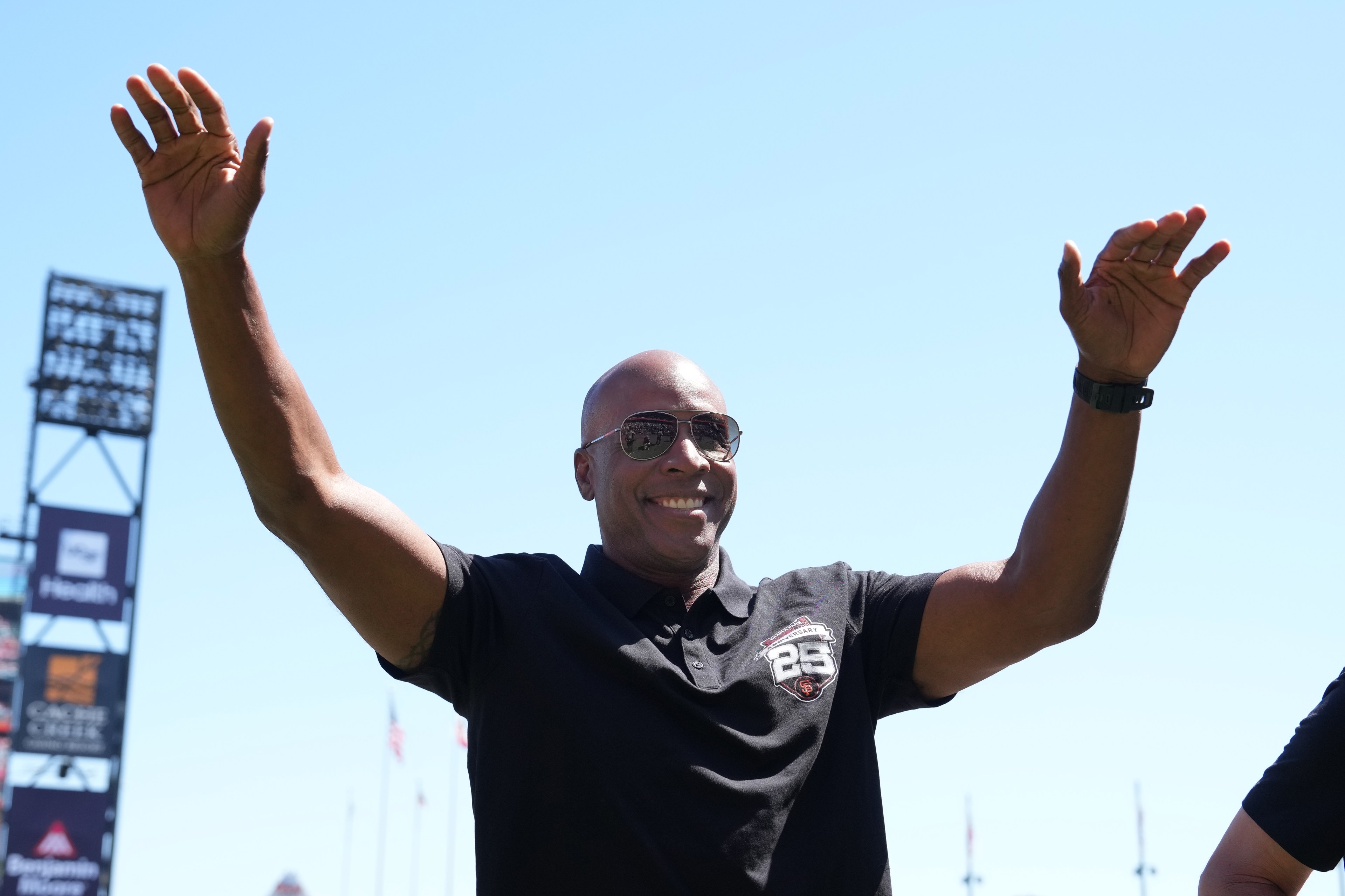 A smiling man in sunglasses and a black shirt raises his arms against a clear blue sky, with stadium signs and flags visible in the background.