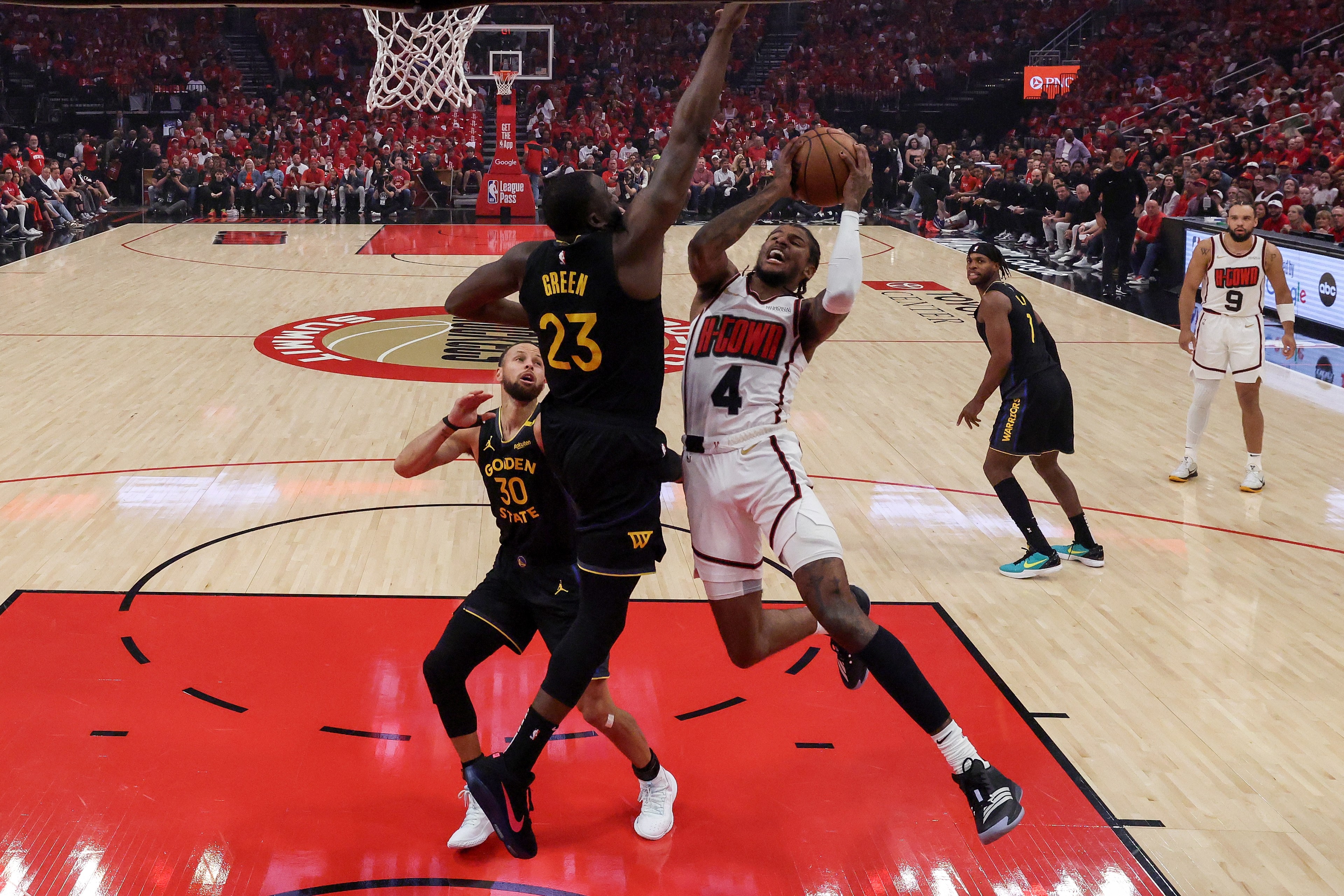 Two basketball players are jumping near the hoop, with one attempting a layup. The court is vibrant and surrounded by an audience dressed in red.
