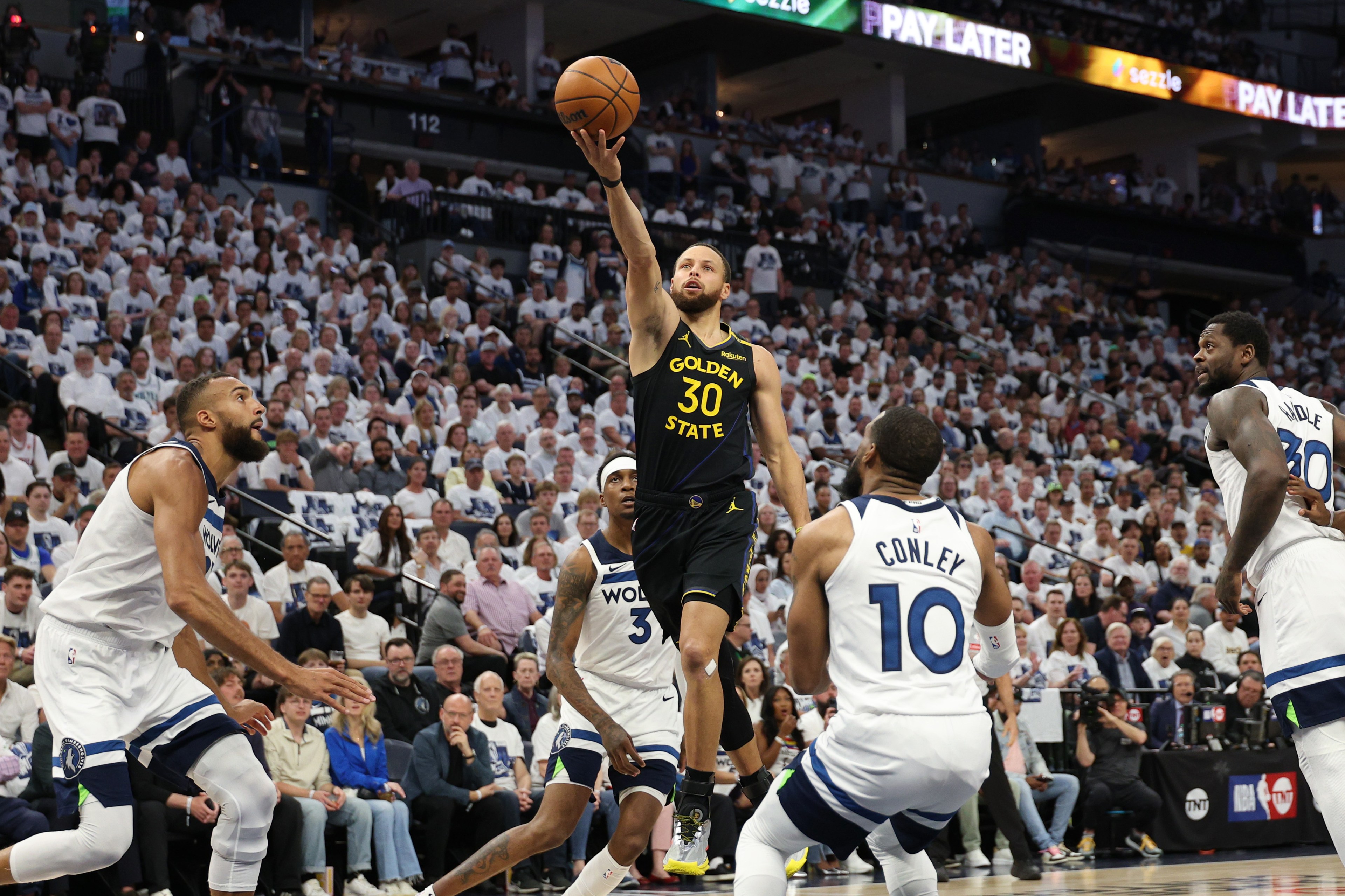 A basketball player in a dark jersey leaps with the ball, aiming for a shot. He's surrounded by four players in white jerseys. The arena is packed with spectators.