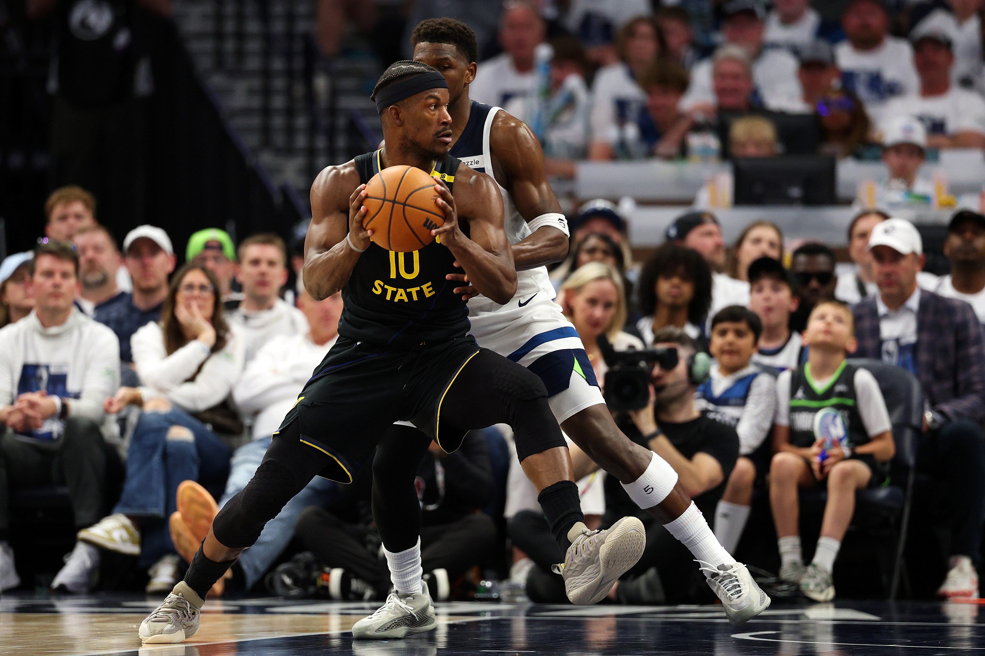 A basketball player in a black jersey holds the ball while another player in white defends. Spectators and photographers watch the action from the sidelines.