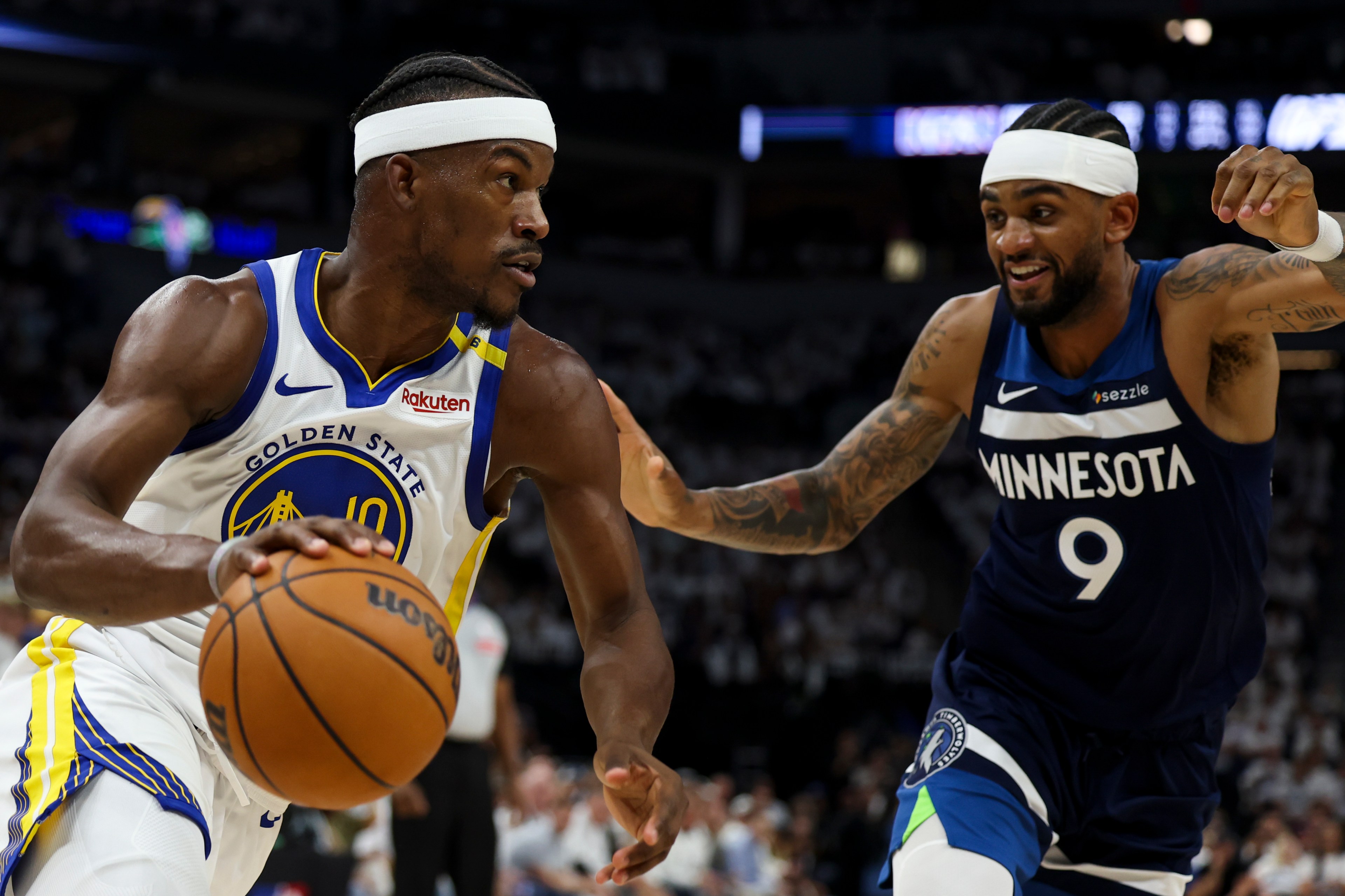 Two basketball players are in action. One in a Golden State jersey dribbles the ball while the other, in a Minnesota jersey, defends. They both wear headbands.