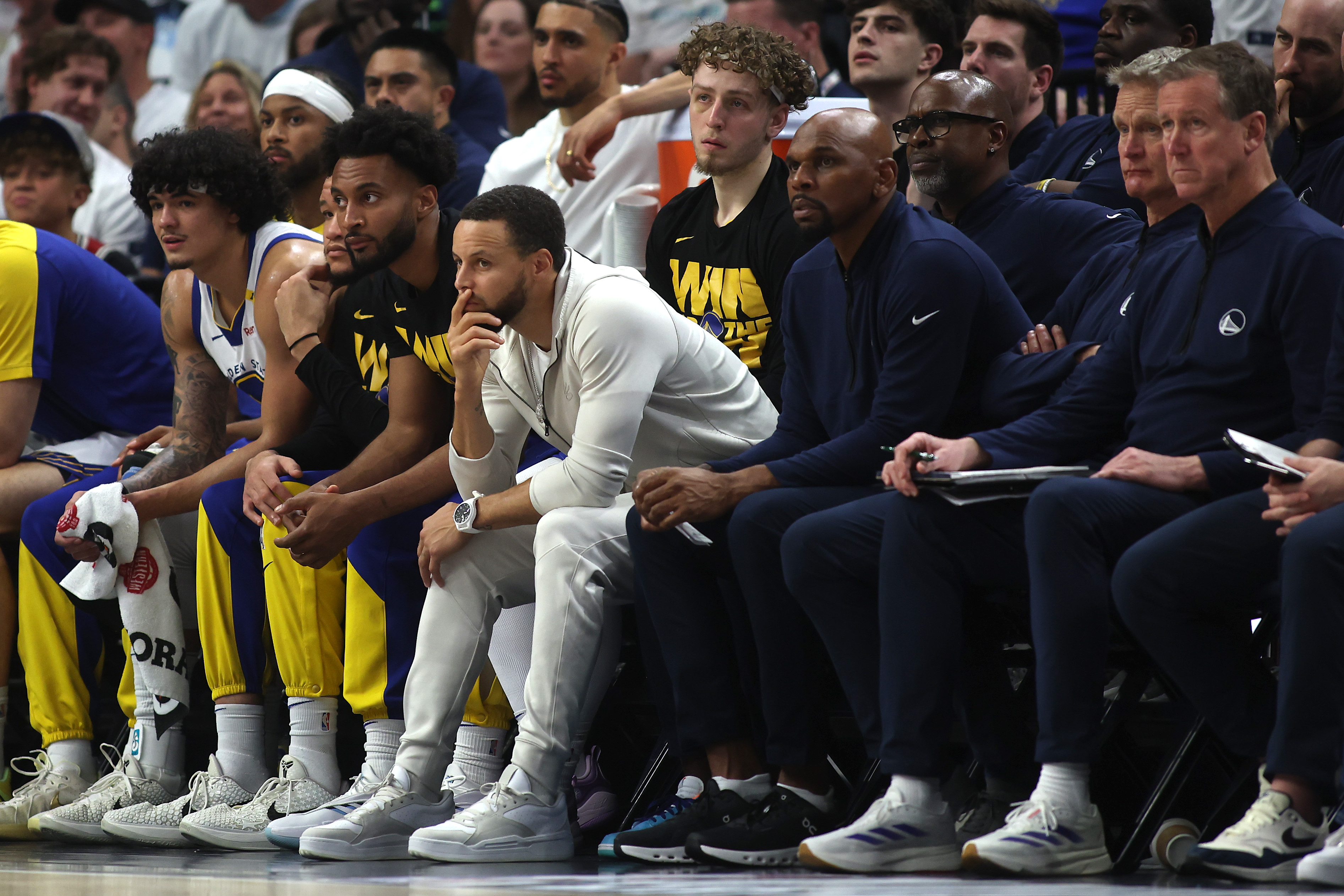 A group of basketball players and coaches sit on a bench. They are focused on the game, with some in team jerseys and others in casual clothes, watching intently.