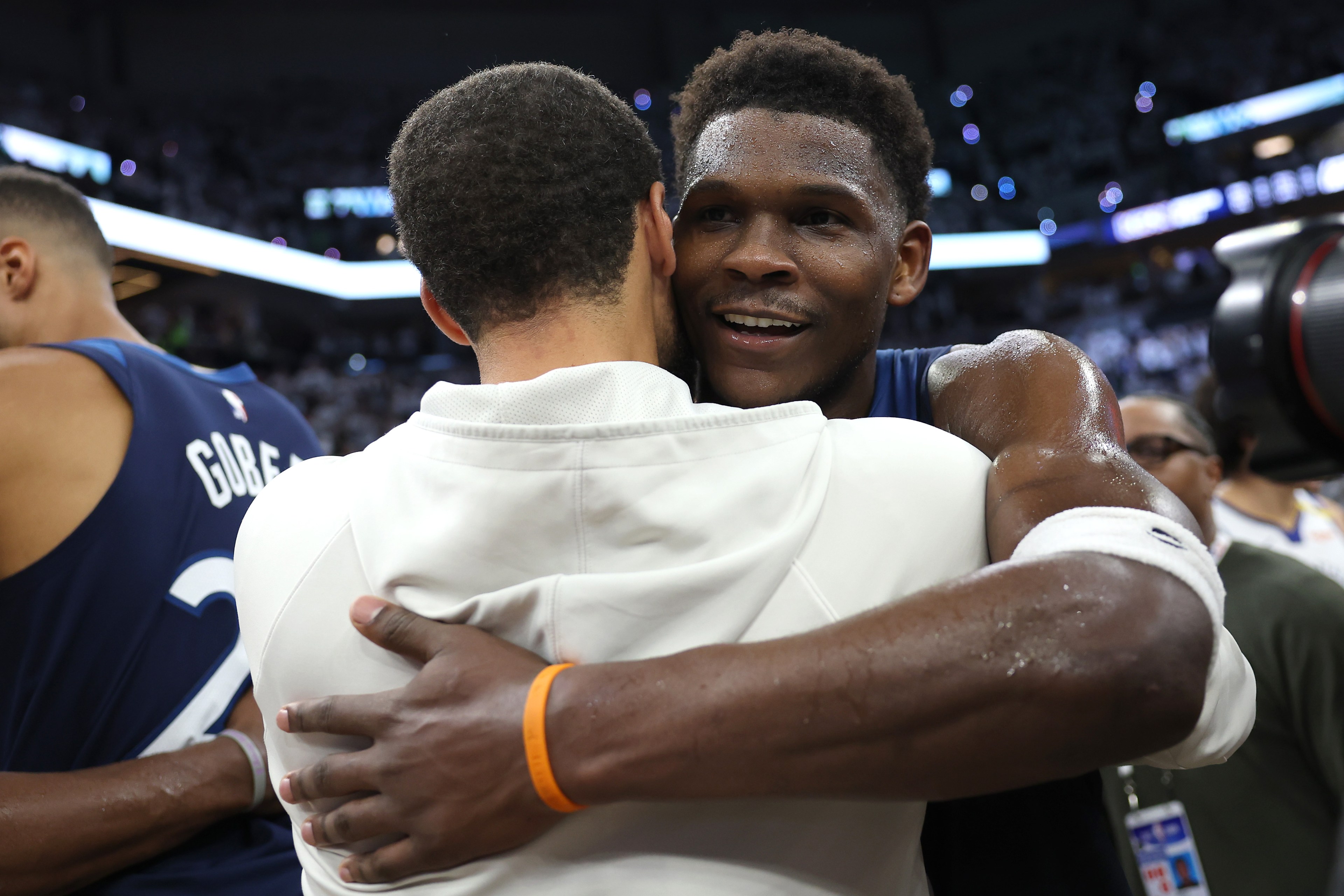 Two basketball players embrace on a court. One player is smiling, sweaty, and wearing a blue jersey, while the other is in a white hoodie, facing away.