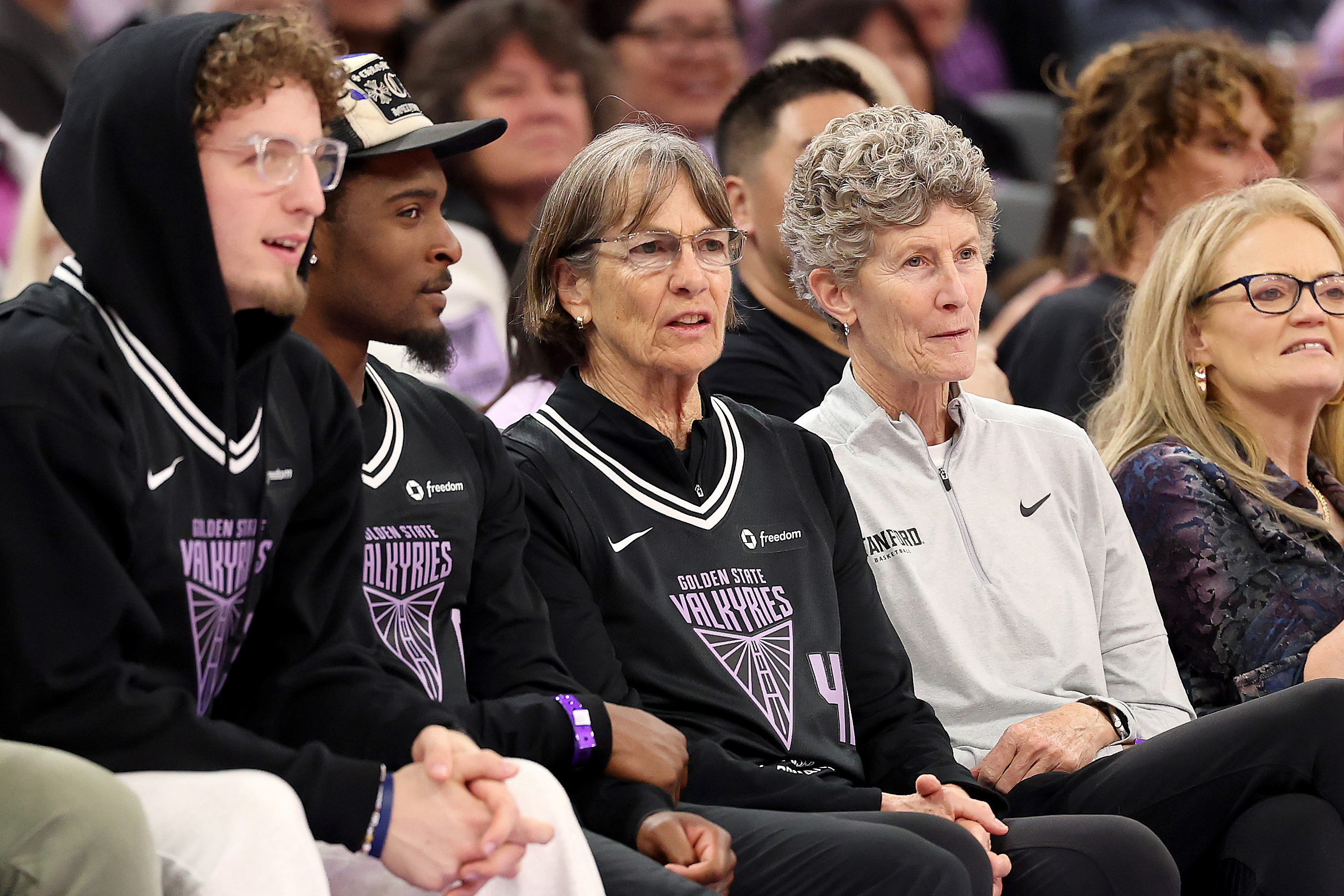 A group of people is seated in a row, mostly wearing &quot;Golden State Valkyries&quot; jerseys. They appear to be watching an event or game, focused and engaged.