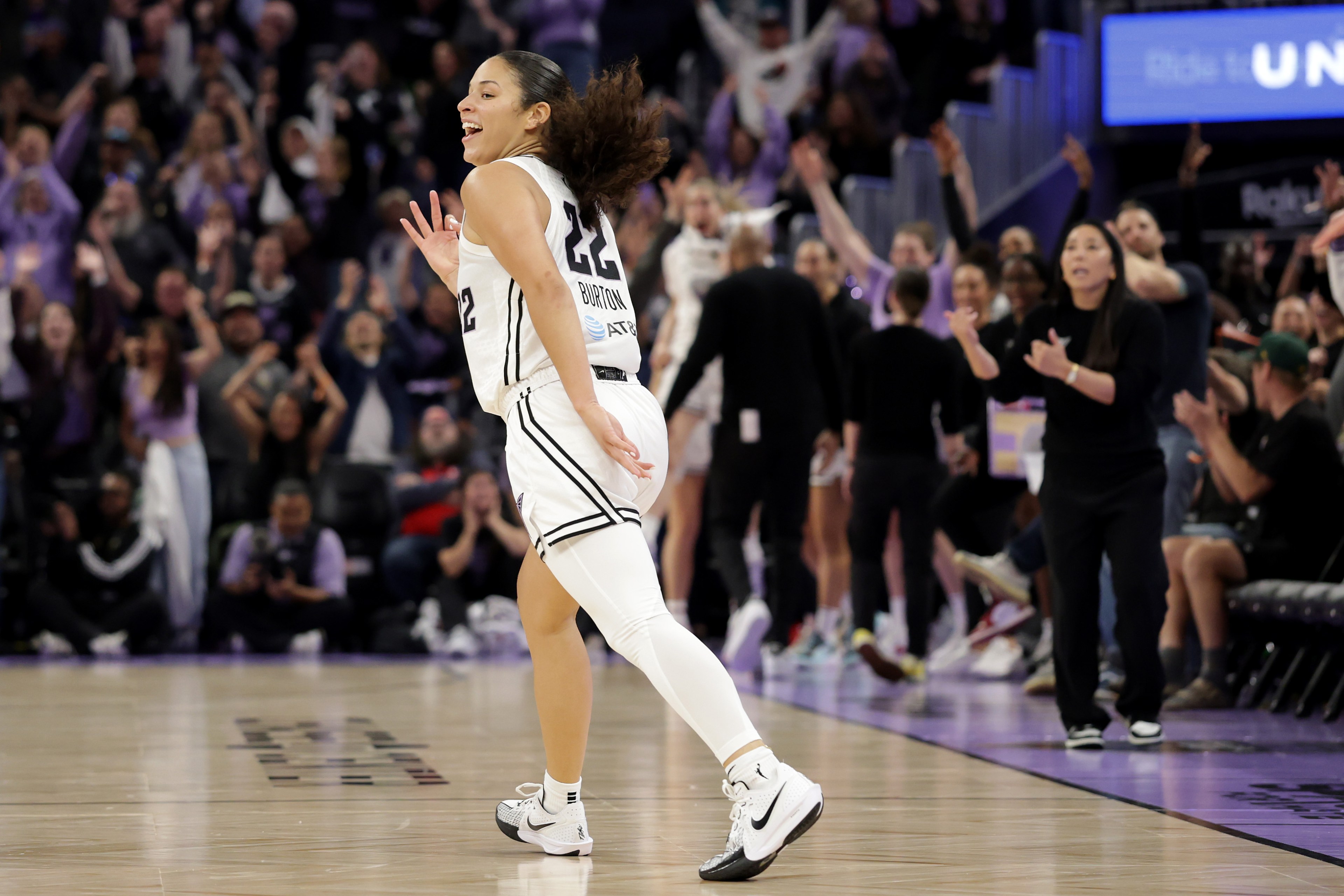 A basketball player in a white uniform celebrates on the court, smiling. The audience and team staff in the background are cheering and applauding.