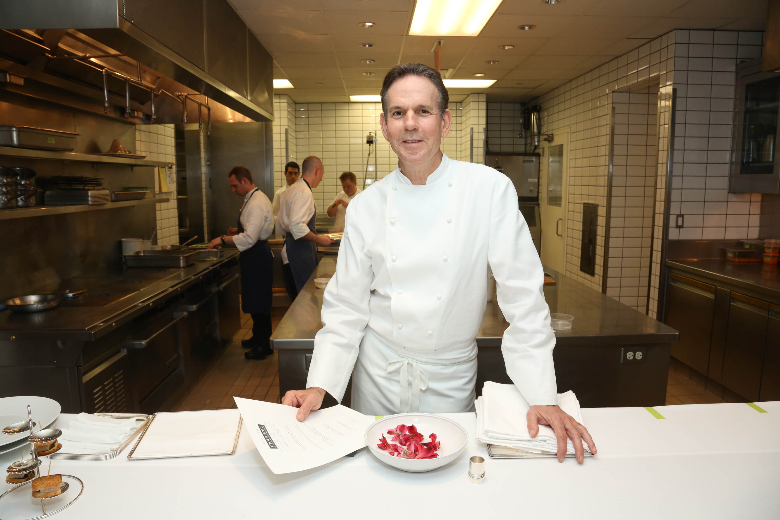 A chef in a white uniform stands in a spotless, tiled kitchen. He holds a paper, with a bowl of red petals nearby. Behind him, others work in the kitchen.