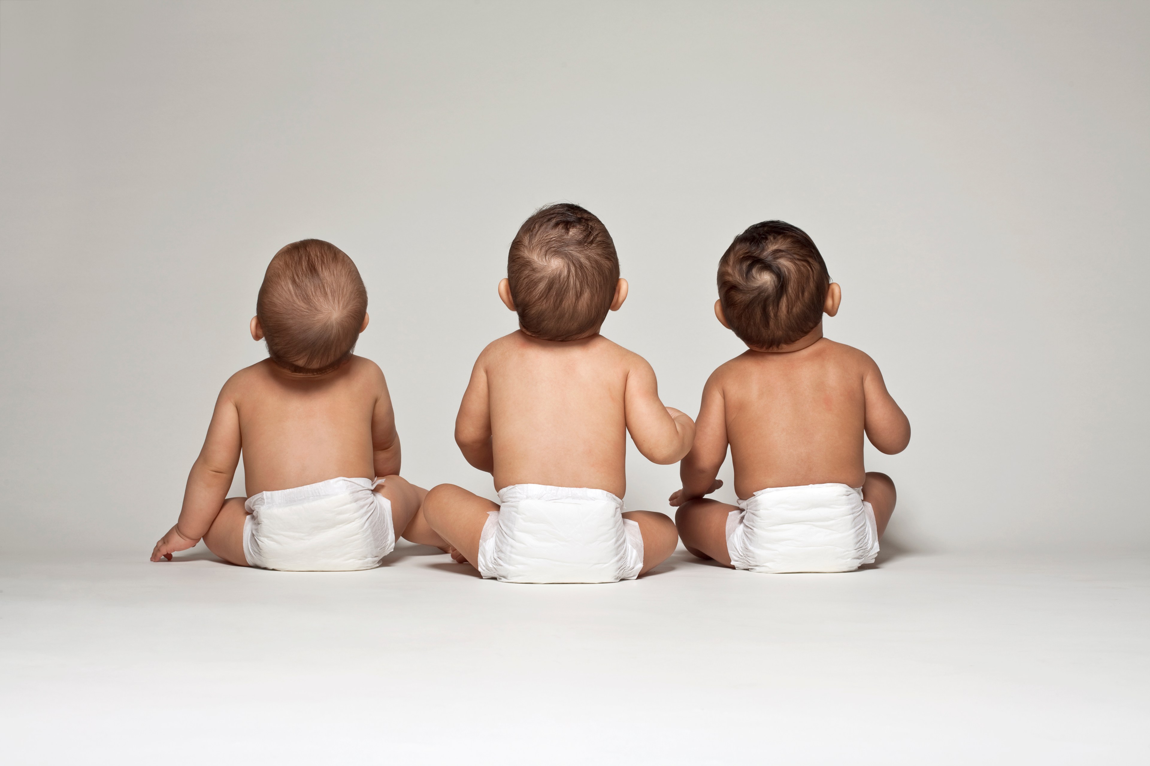 Three babies in diapers sit side by side, viewed from behind, on a plain white background. The central baby has their hand on the shoulder of another.