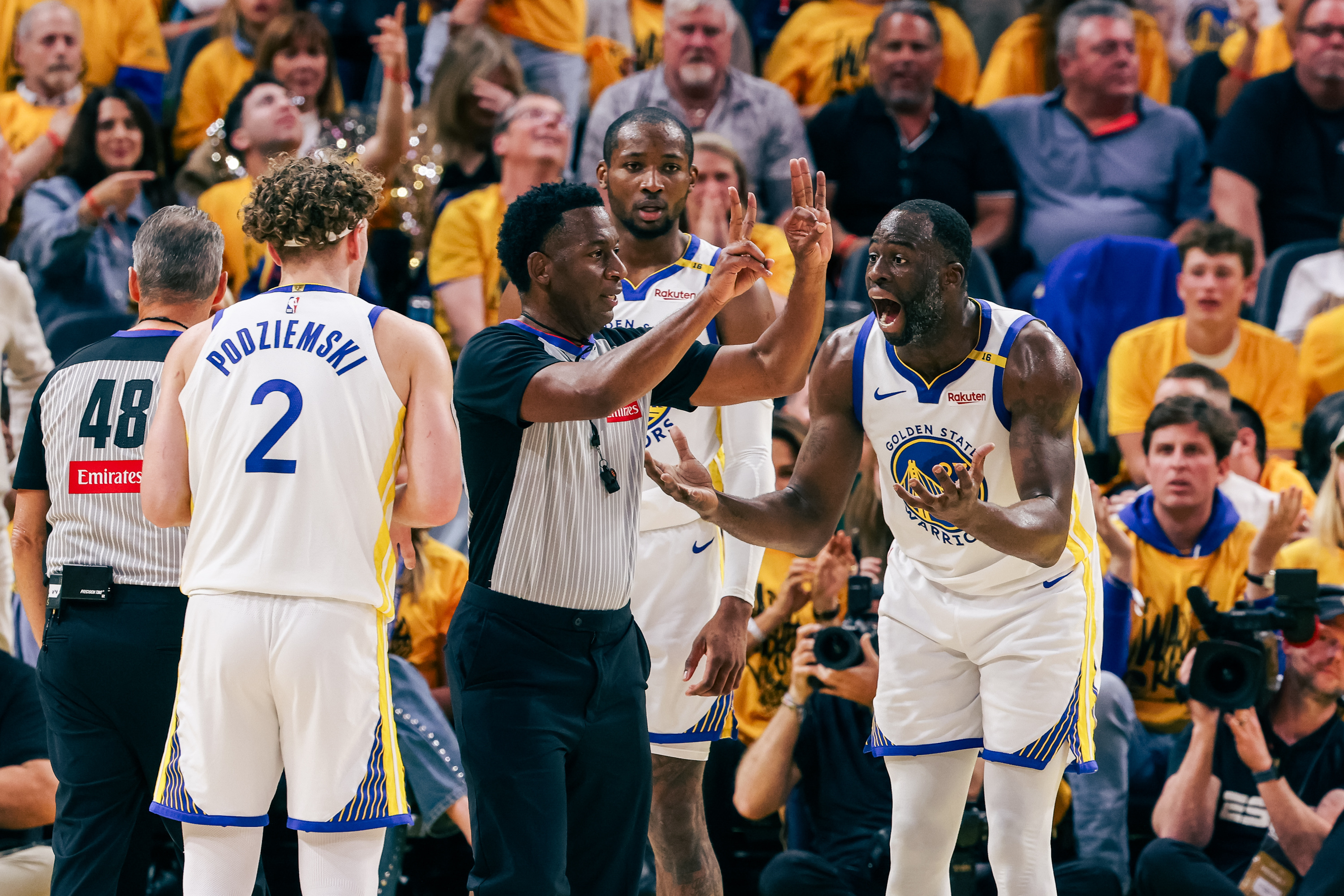 A basketball player in a Golden State Warriors jersey argues with a referee on the court. Another player and a referee are nearby, and fans are in the background.