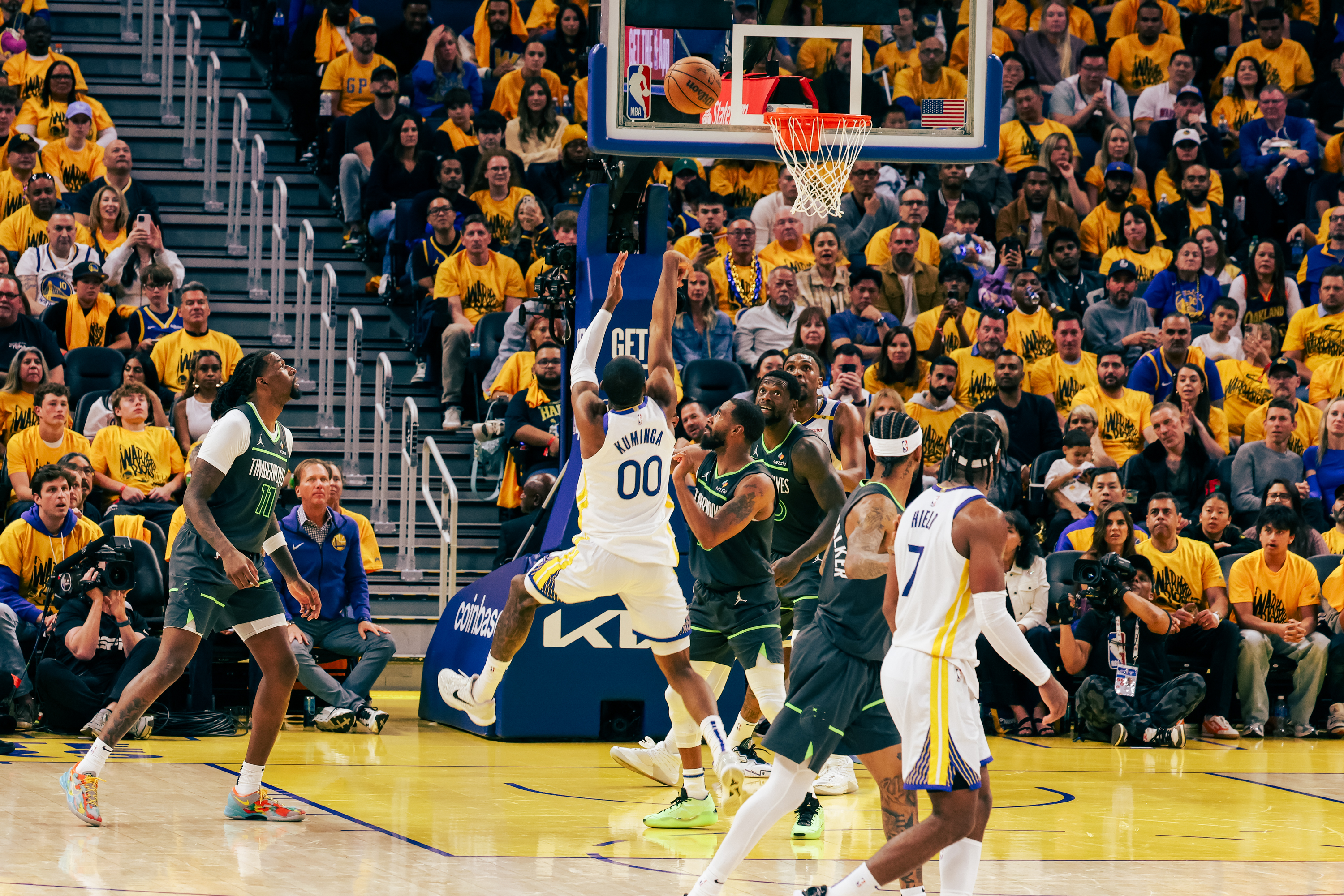A basketball player in a white jersey jumps to score as players in green defend. The court is vibrant with fans in yellow cheering energetically in the stands.