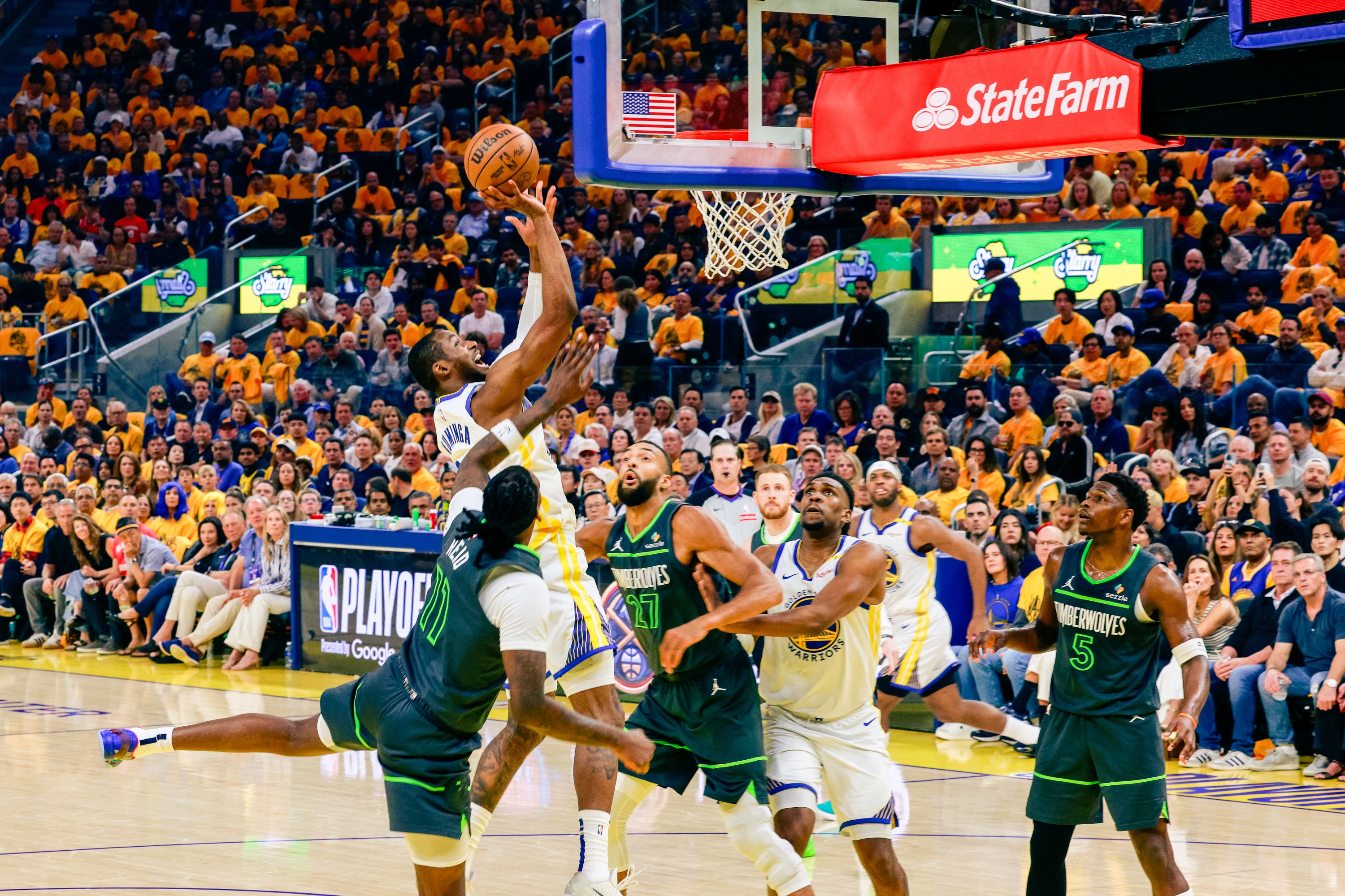 A basketball player in a white and blue uniform leaps toward the hoop to score, surrounded by opposing players in green. The crowd watches intently.