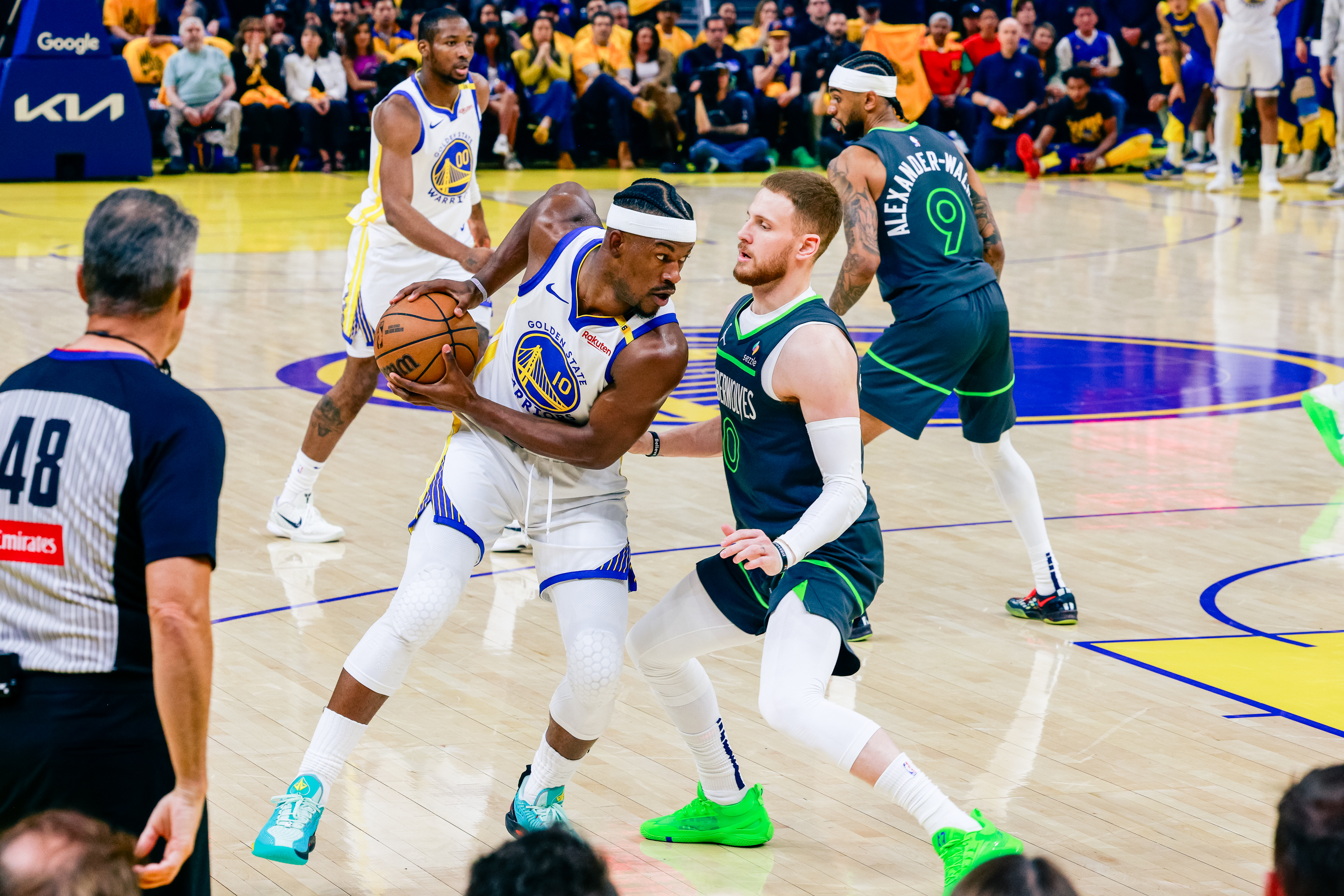 A basketball player in a white uniform is holding the ball, guarded by a player in a dark green uniform. A referee and other players are visible in the background.