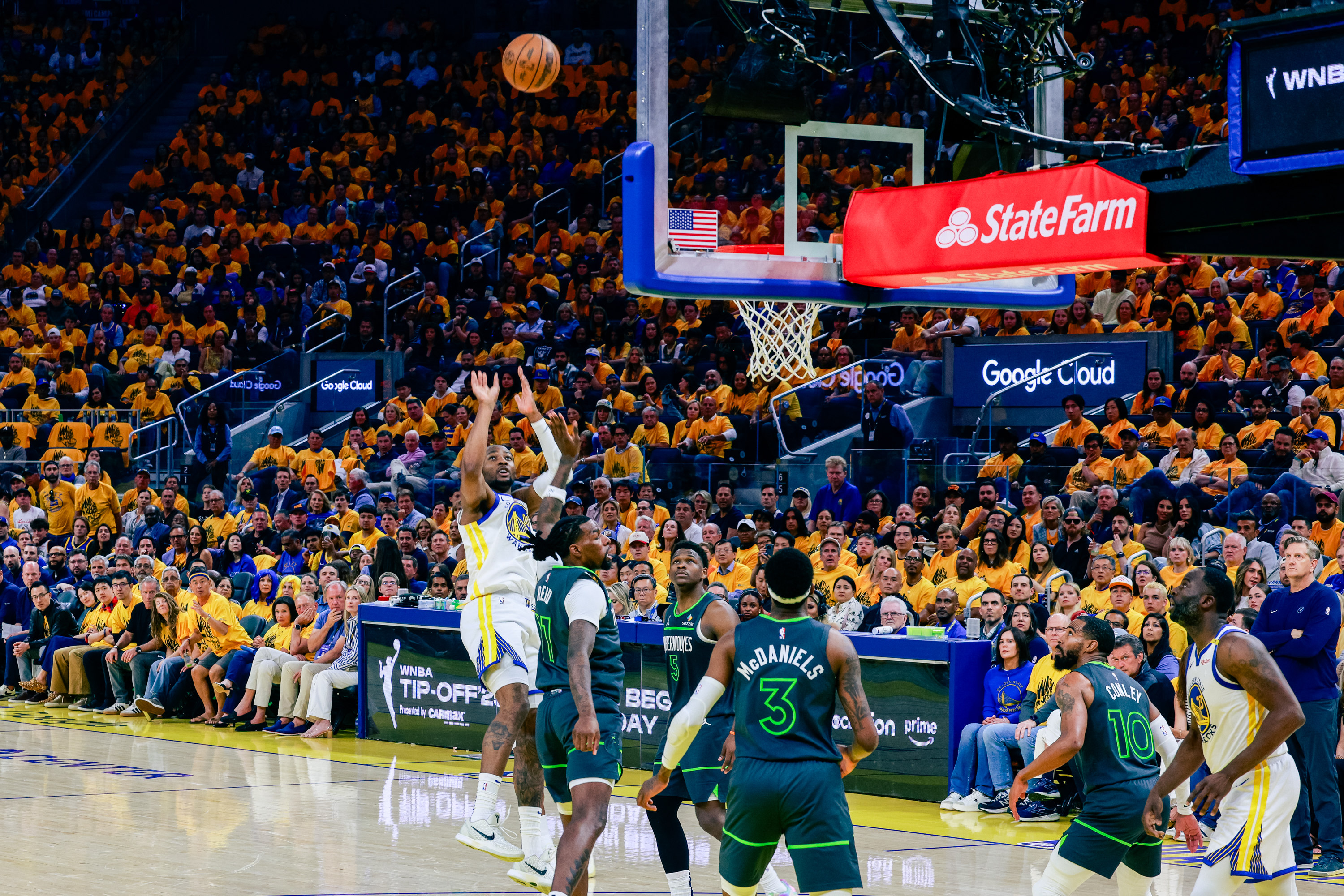A basketball game is in progress with a player in a white and yellow jersey attempting a shot, while players in dark jerseys watch. The audience is vibrant.