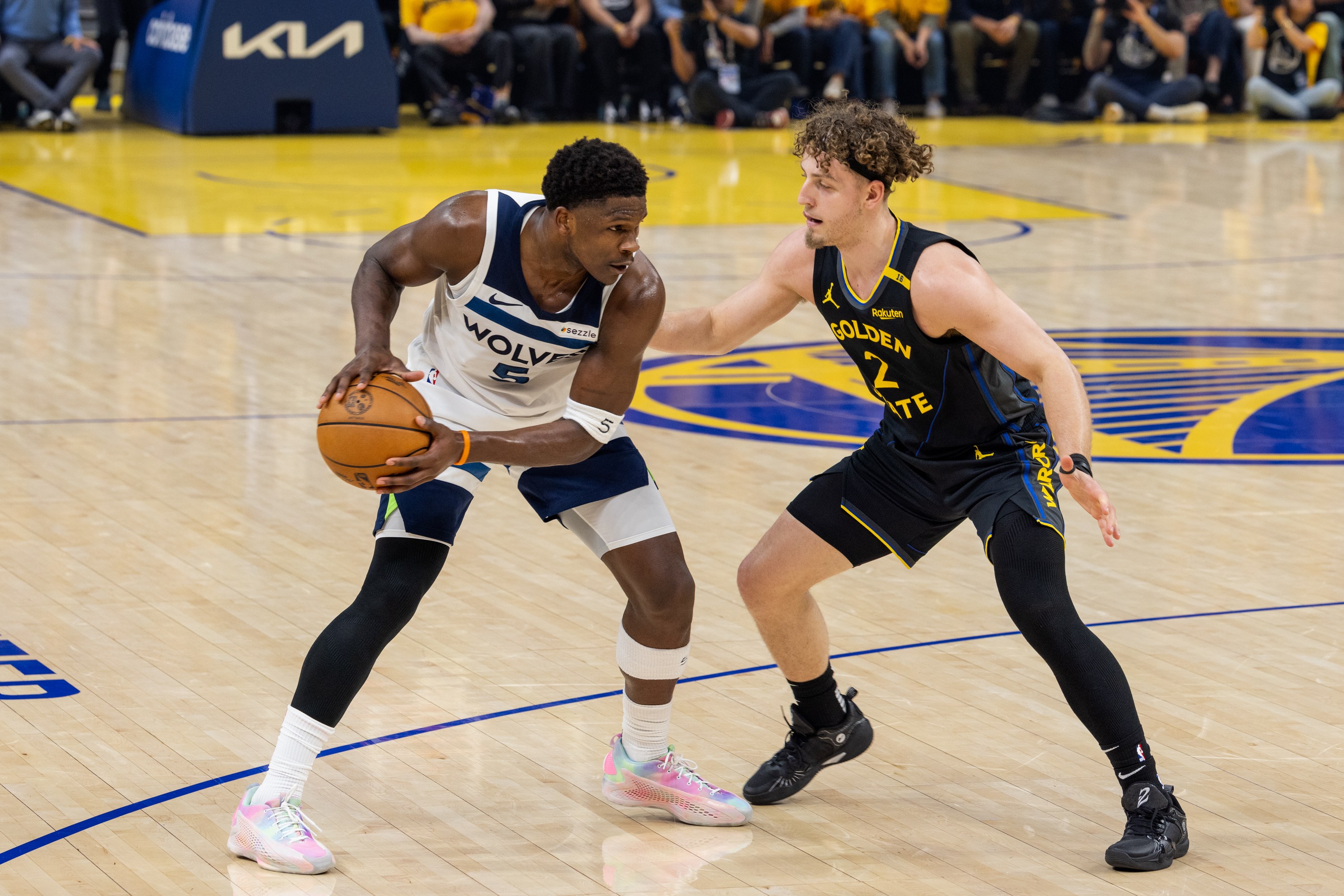 A basketball player in a Wolves jersey holds the ball, guarded closely by a player in a Golden State uniform on a court.
