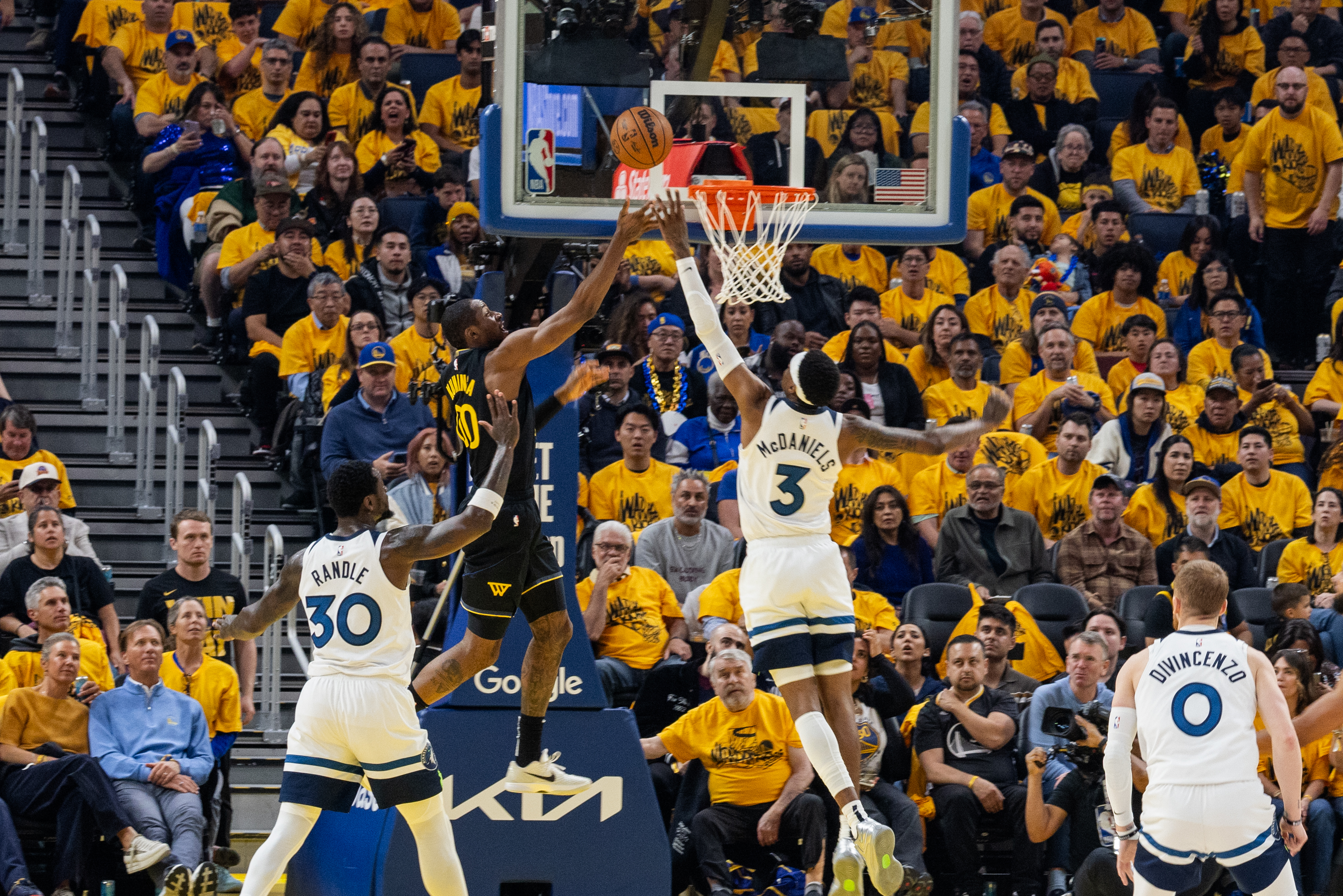 Two basketball players in black and white jerseys jump for a rebound near the hoop, with a crowd of fans wearing yellow shirts in the background.