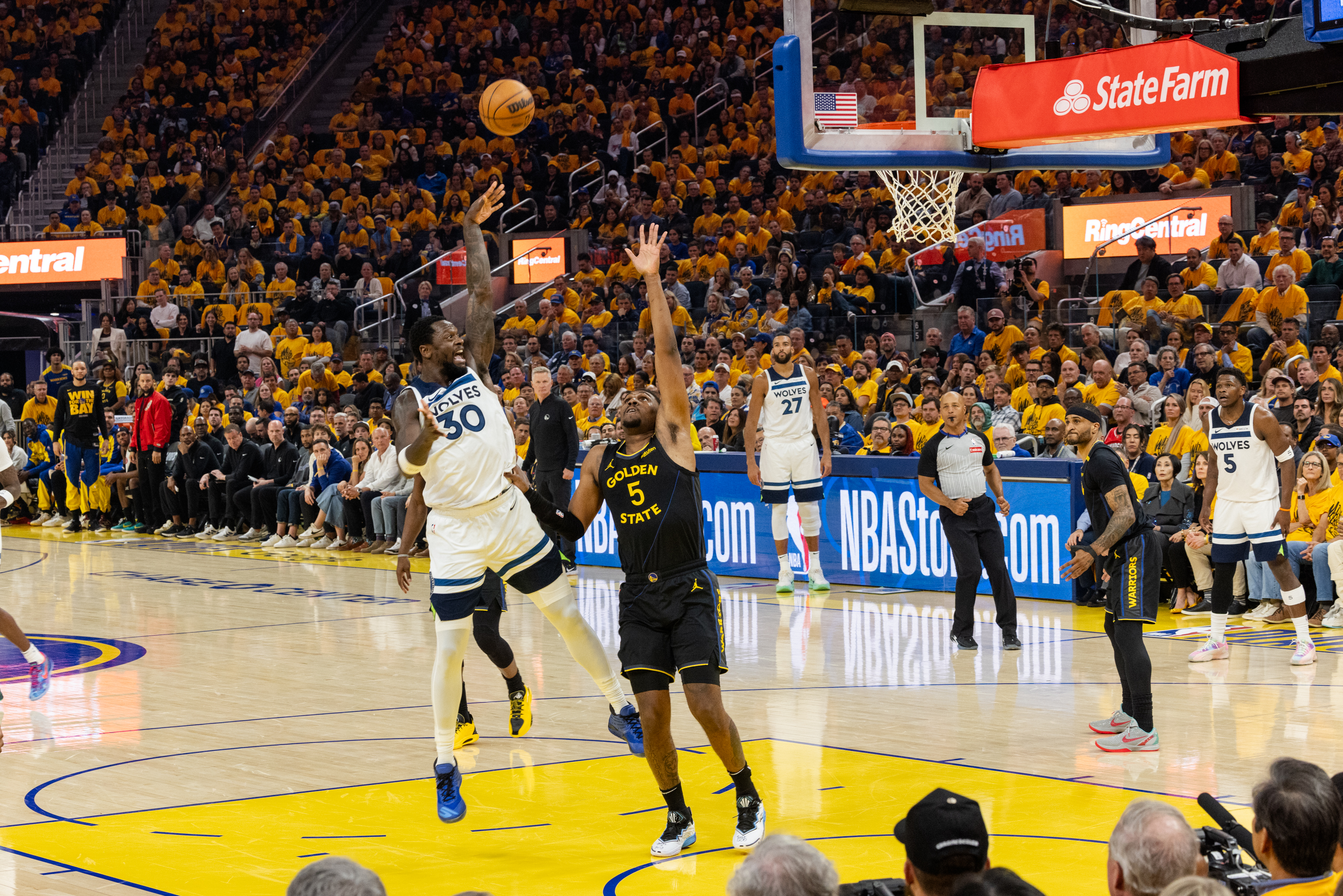A basketball player in a white jersey jumps to shoot the ball while a player in a black jersey defends. An audience in yellow shirts watches intently.