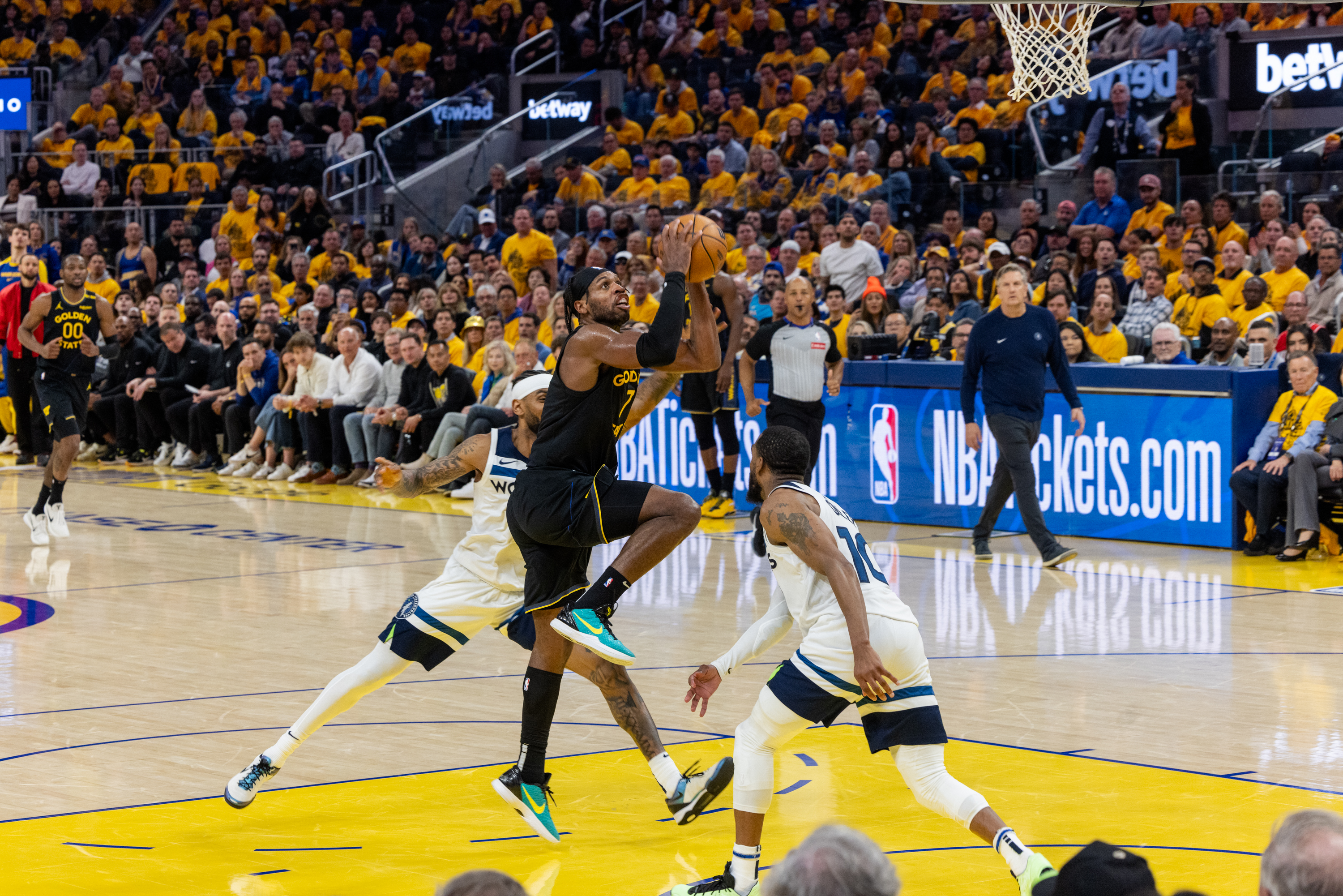 A basketball player in a black jersey is jumping with the ball near the hoop. Two players in white jerseys defend him, while the audience watches intently.