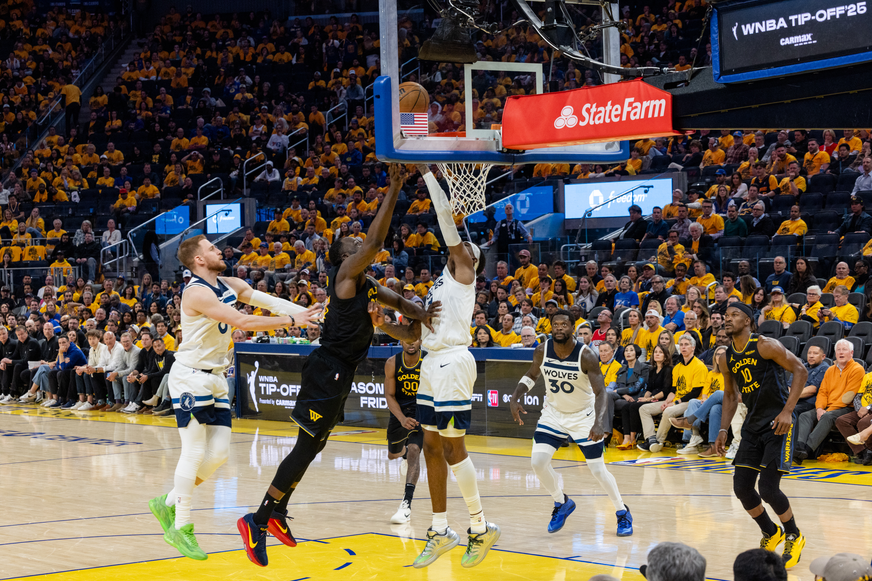 Players from two basketball teams are competing under a hoop. One player in black attempts a block, while others in white and black jerseys are around. The crowd watches intently.
