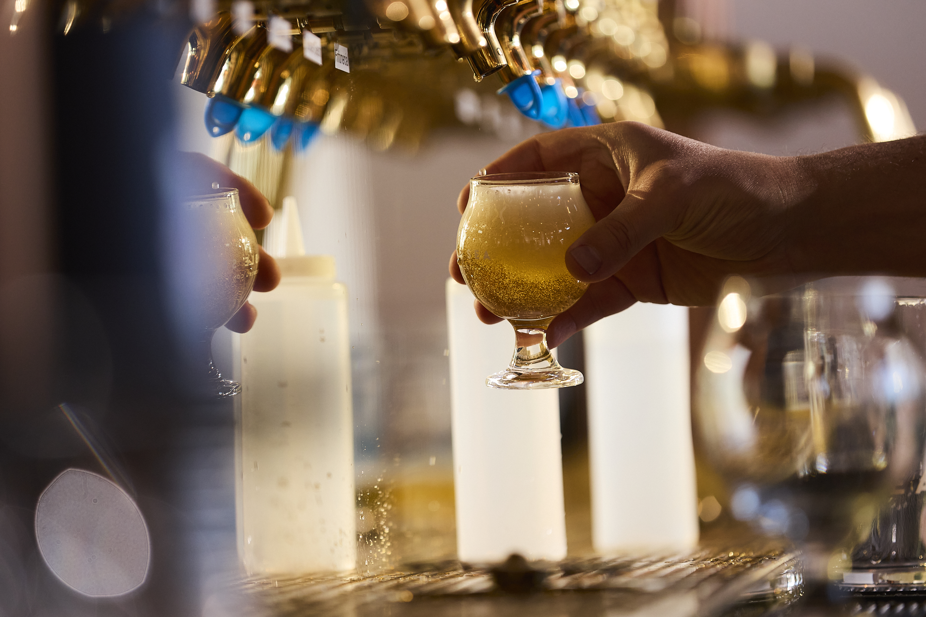 A hand holds a small, filled beer glass with a foamy top under golden taps. The background includes white bottles and a blurred effect with light reflections.