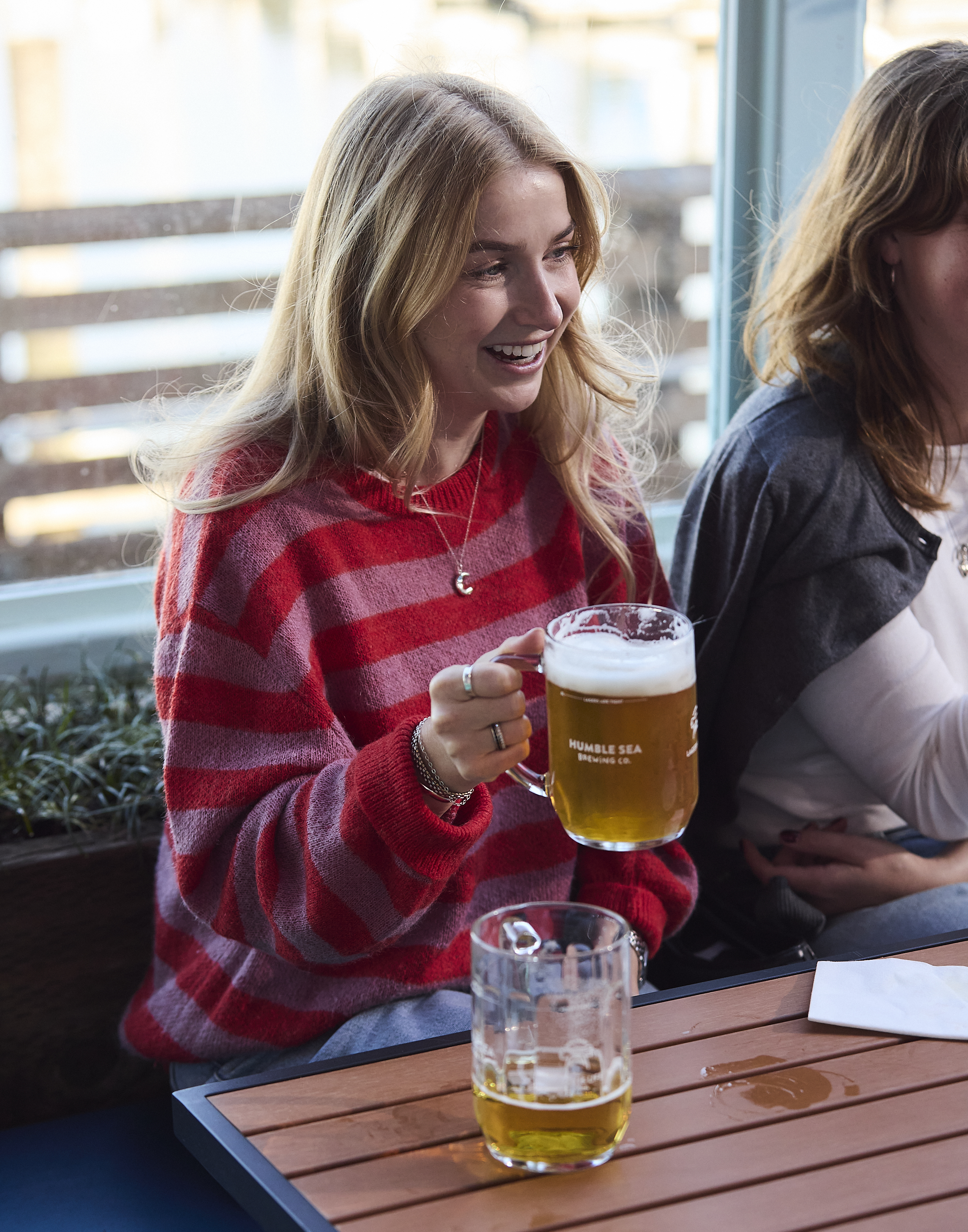 A smiling person with long hair, wearing a red striped sweater, holds a beer mug while sitting at a wooden table beside another person.