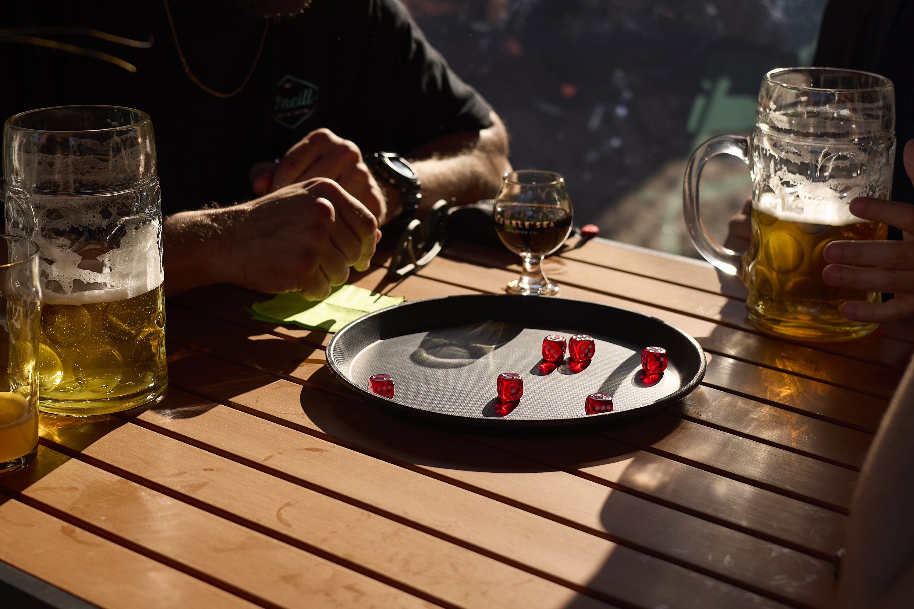 Two people sit at a wooden table with two large beer mugs, a small glass, a tray holding red dice, and sunlight casting shadows.