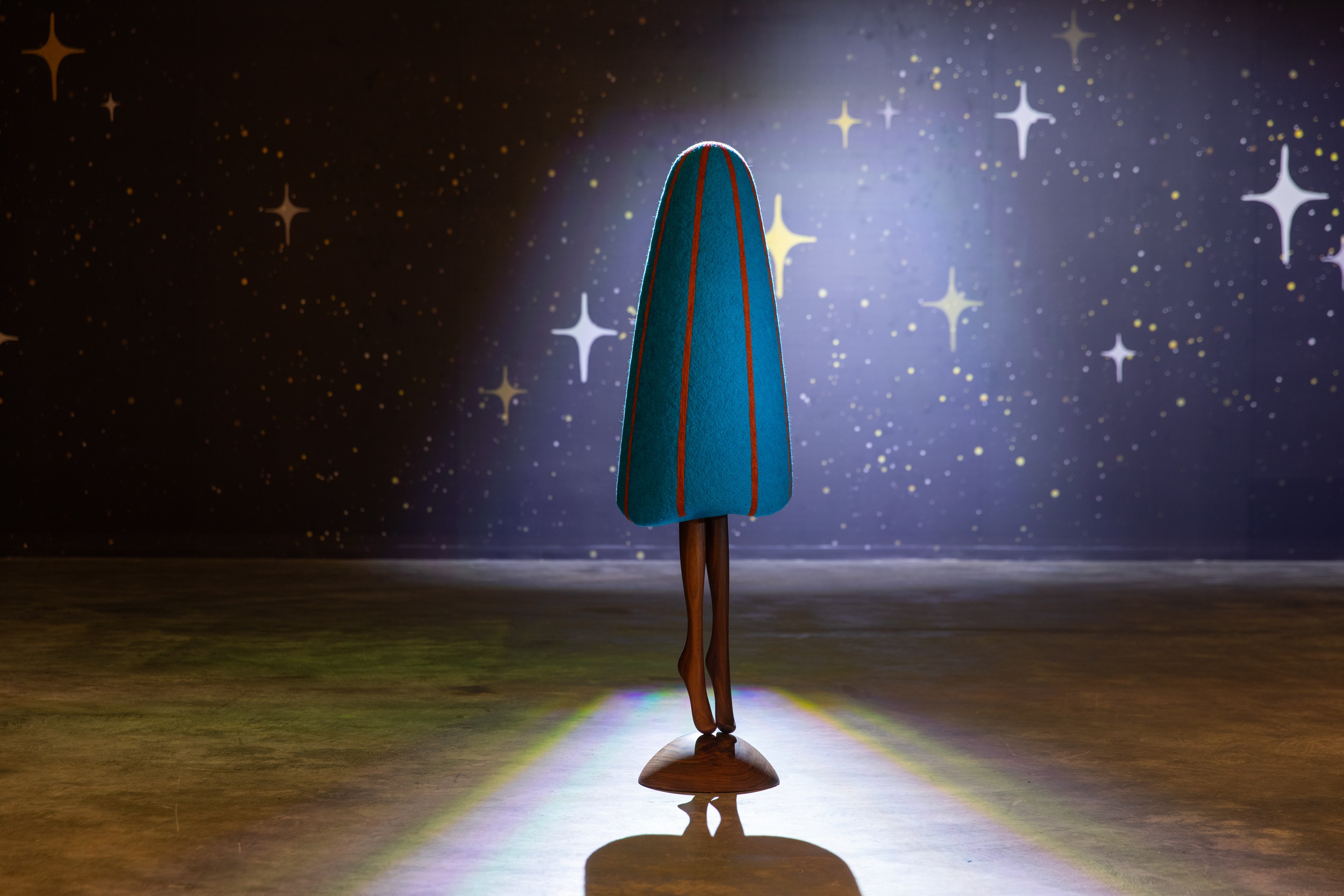 A wooden sculpture with raised legs stands on a cone-shaped base. It's covered by a blue felt fabric with red stripes, set against a starry backdrop.