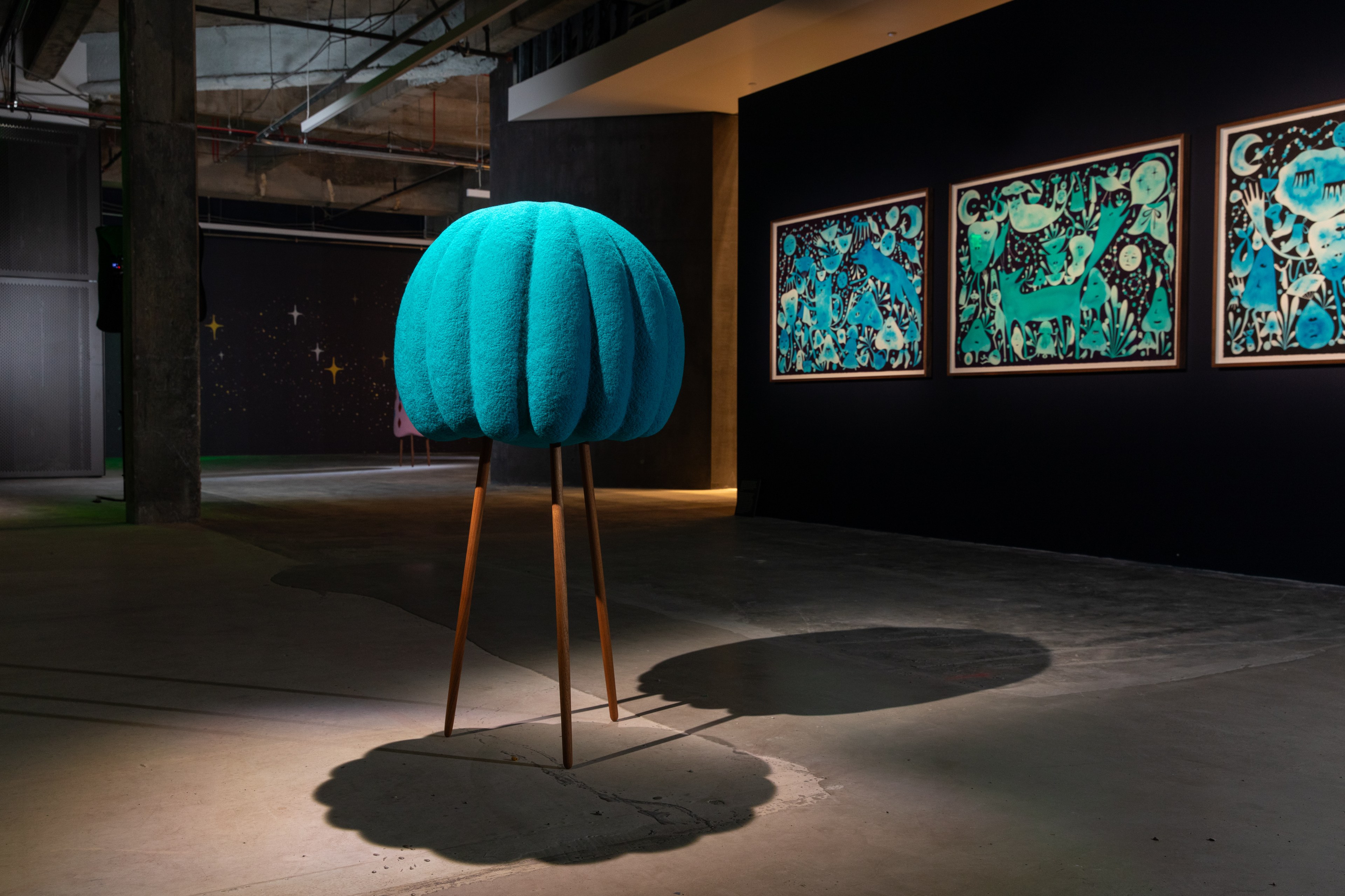 The image shows a cyan, textured sculpture on wooden legs, resembling a lollipop. It stands in an art gallery with three abstract paintings on the wall.