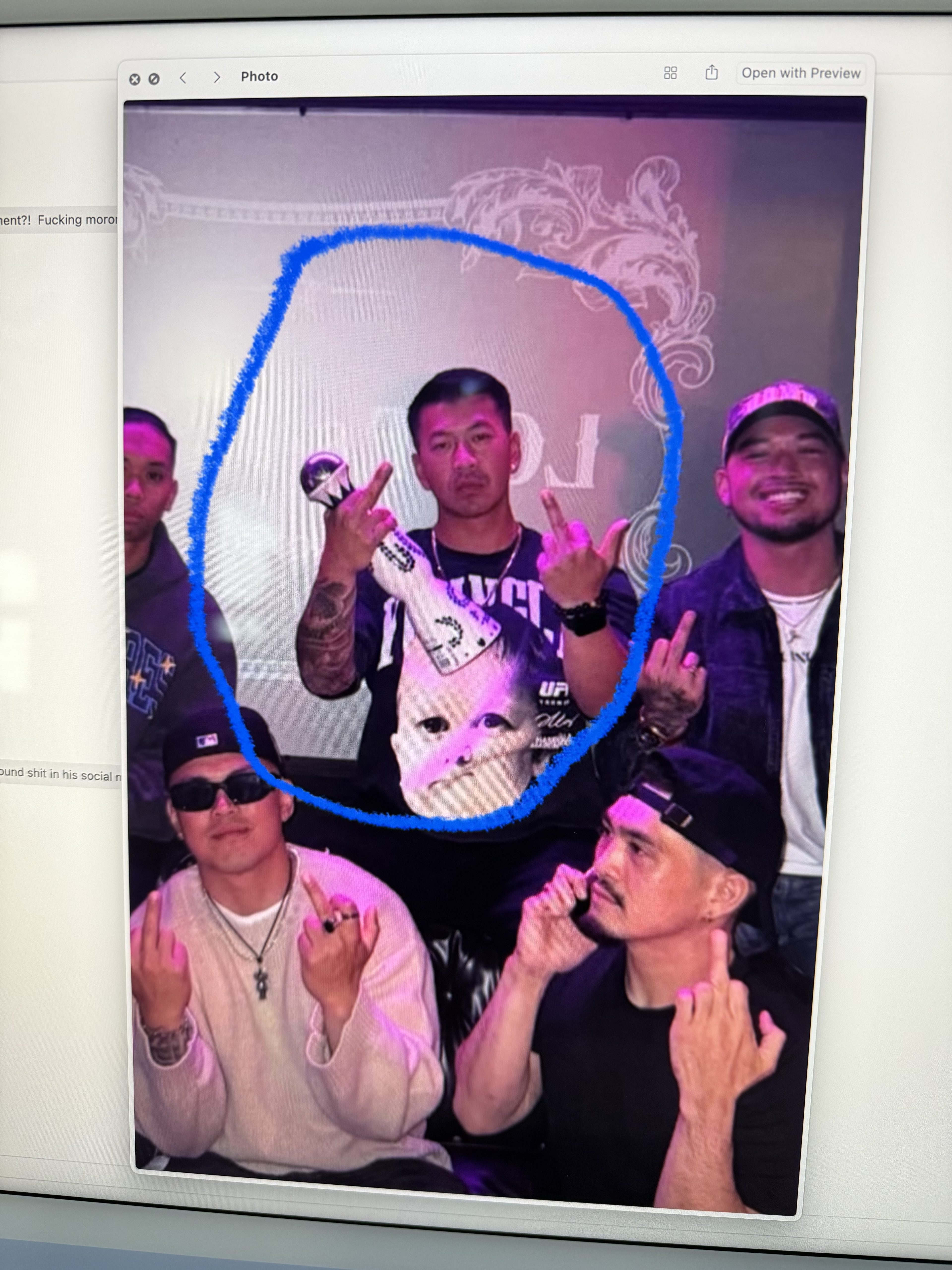 Five men are posing, making rude gestures. One in the center holds a statue, wearing a shirt with a baby face. The atmosphere is casual with bright lighting.