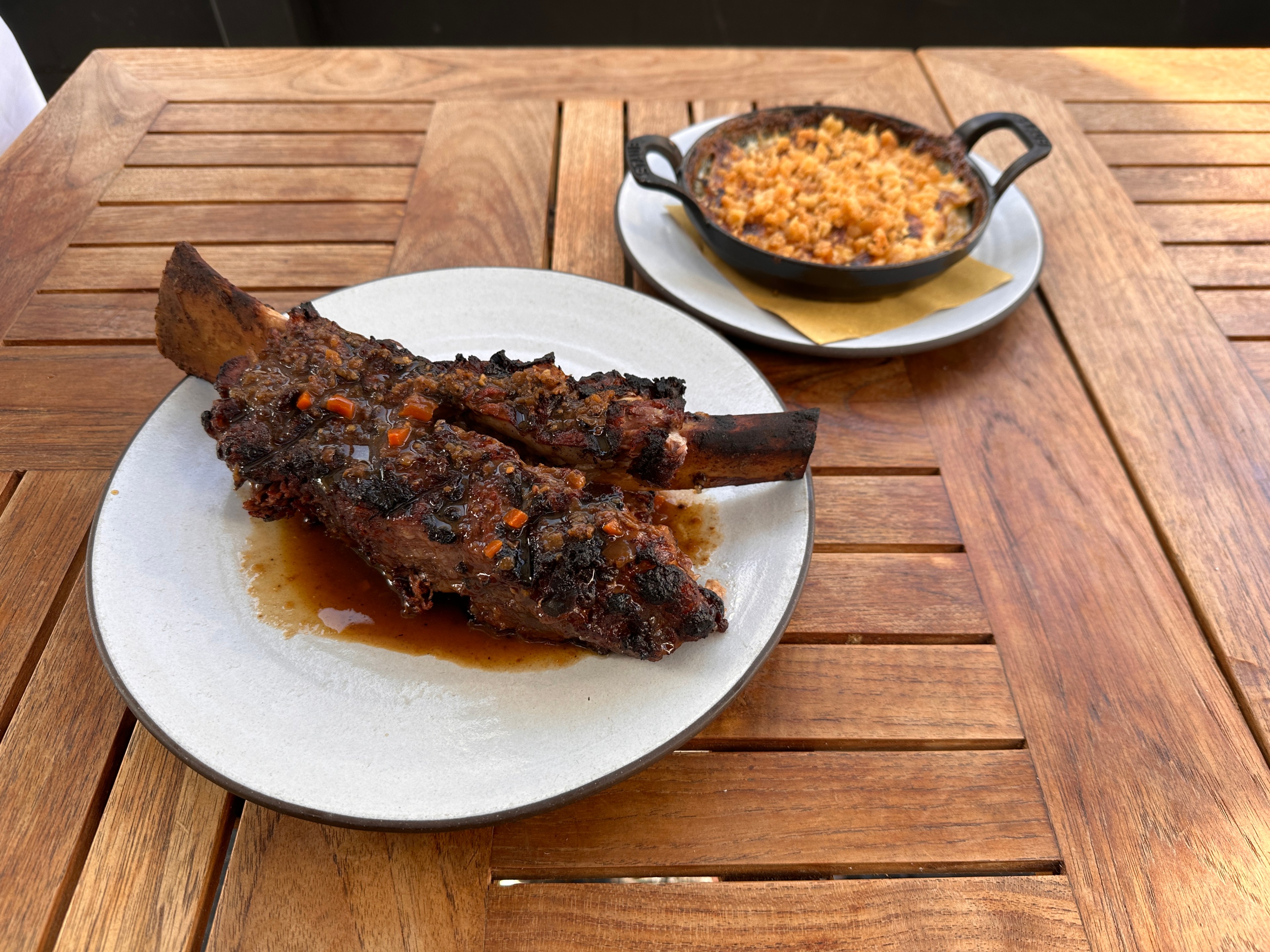A wooden table holds a plate of juicy, glazed ribs with vegetables, next to a small cast iron pot filled with a macaroni and cheese dish.