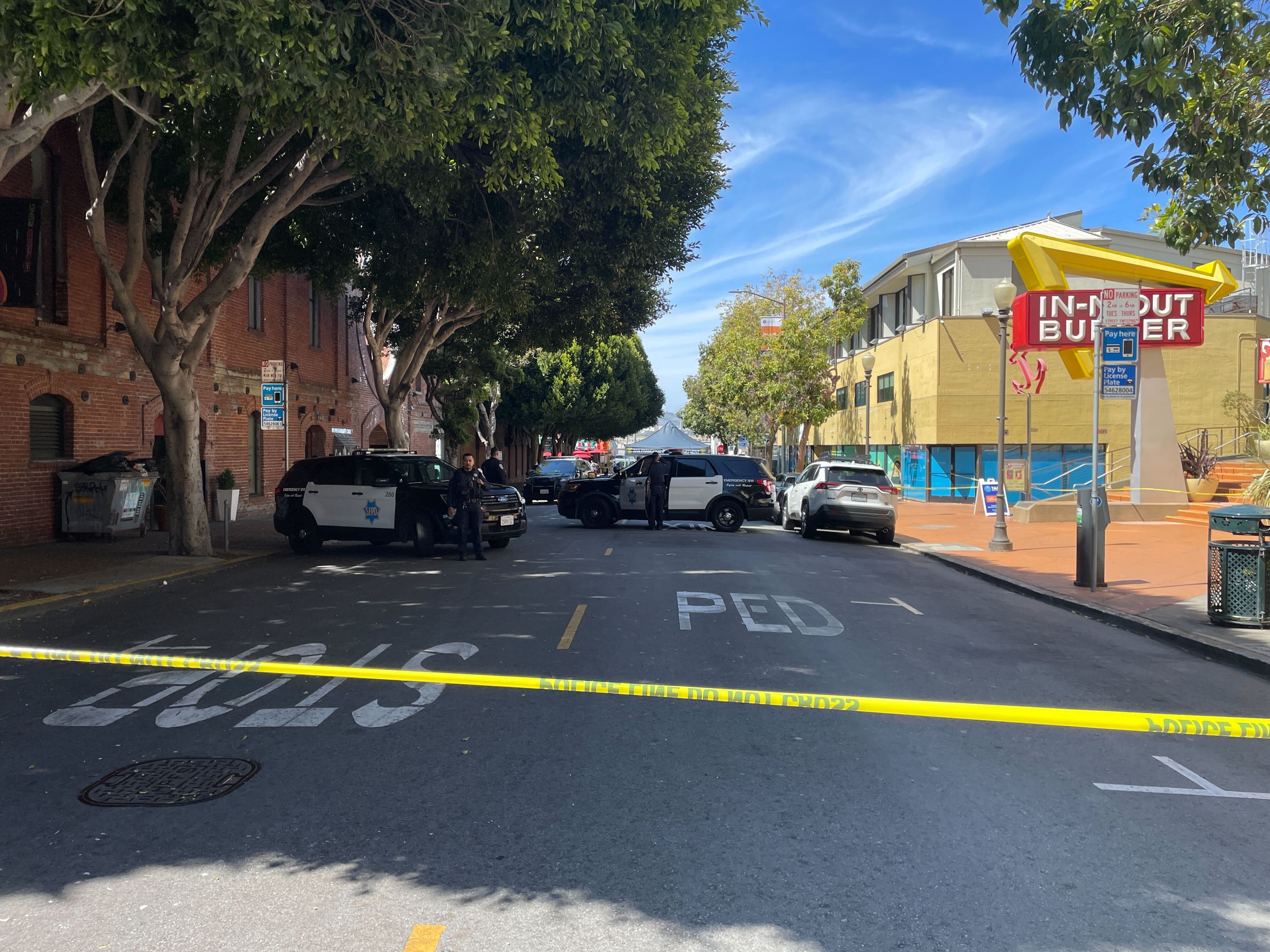 Police cars block a street lined with trees and buildings, including a visible burger restaurant. Yellow tape cordons off the area, and officers are present.