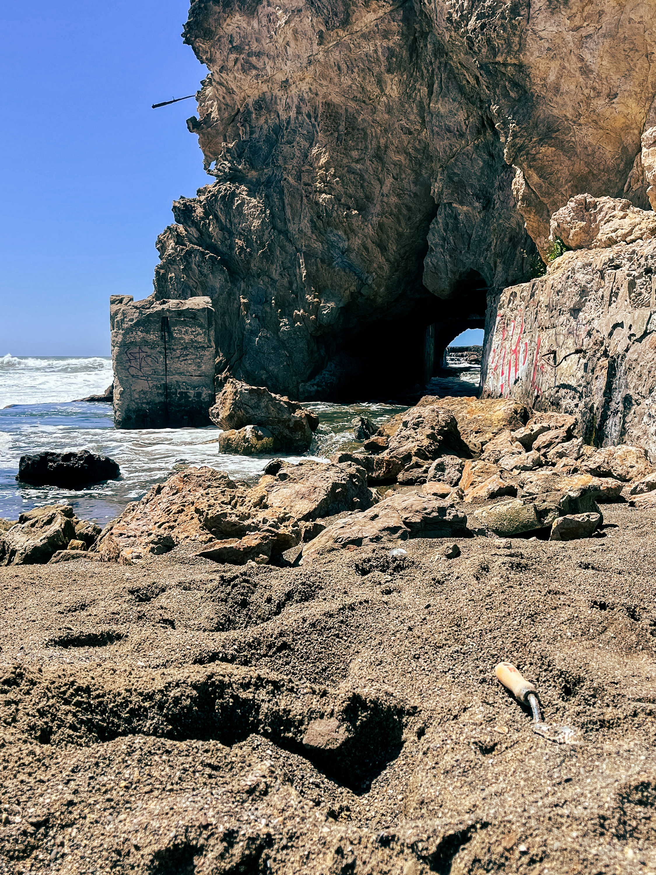 A rocky beach scene with a large cliff and a dark cave. The ocean waves crash against the rocks, and there's a small trowel lying on the sandy ground.