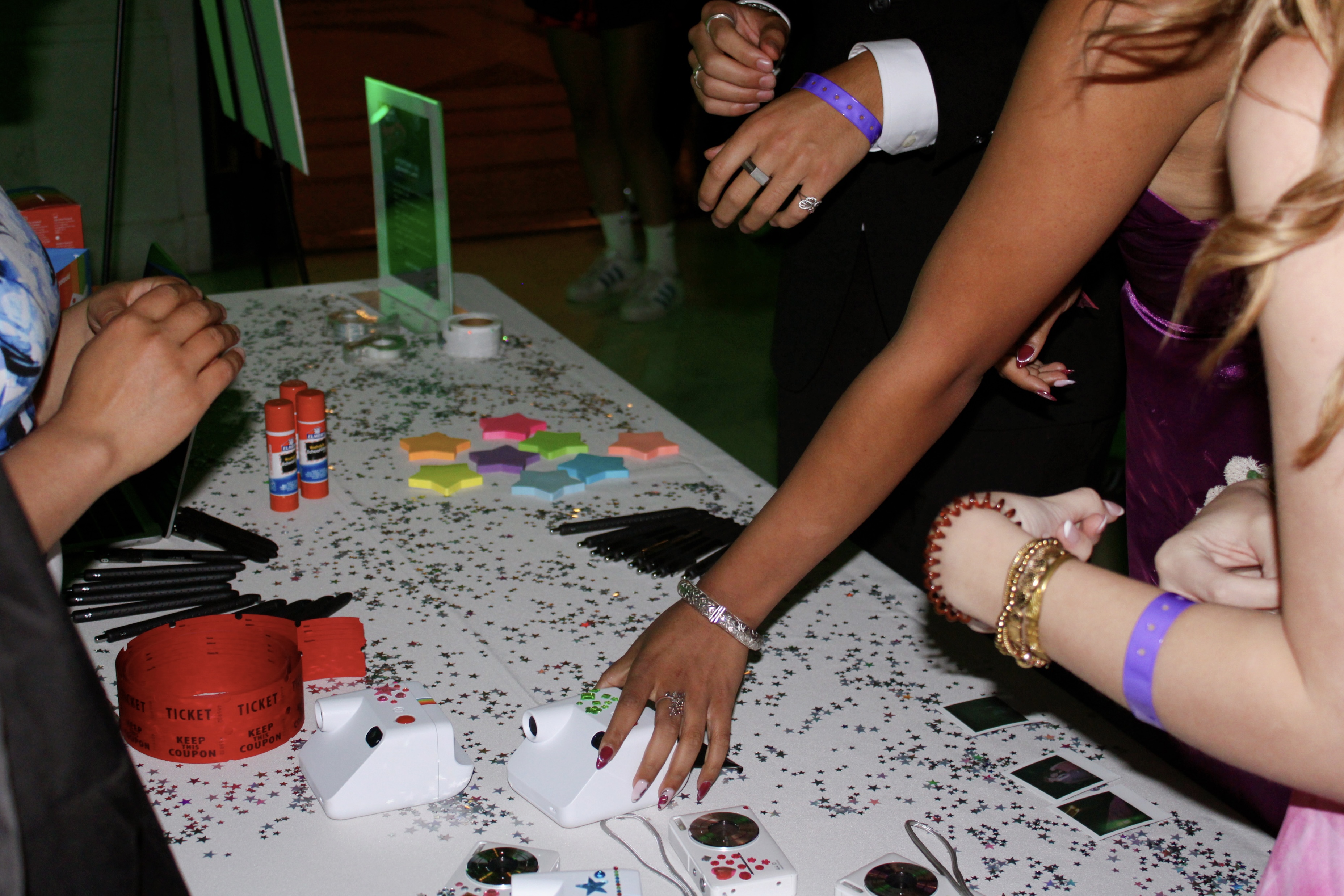 A group of people at a table with colorful star-shaped notes, pens, glue sticks, and rolls of tickets. The table is sprinkled with star confetti.
