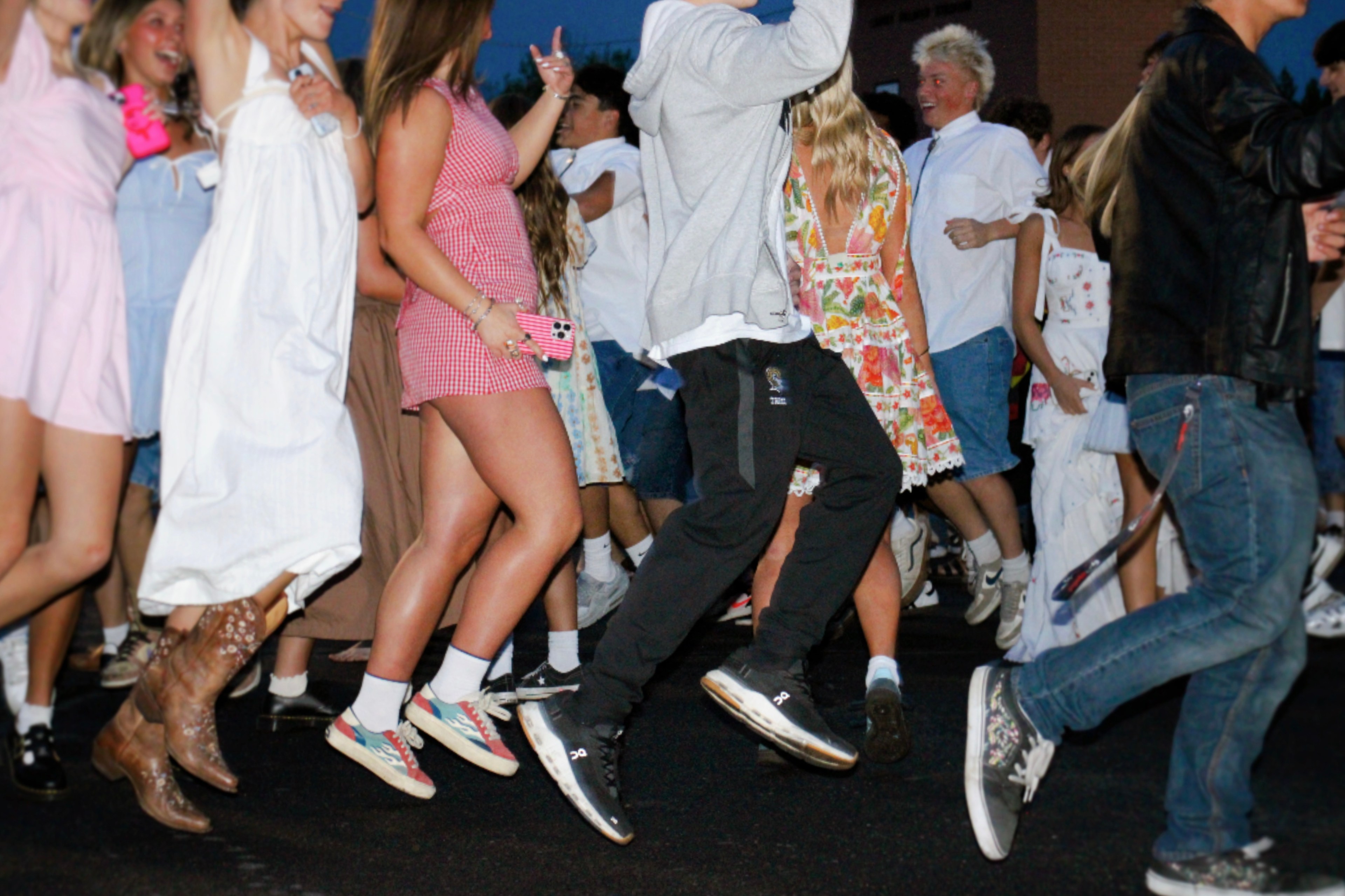 A group of people are energetically dancing outside. They're dressed in casual and summer attire, with some in dresses and sneakers, creating a lively atmosphere.