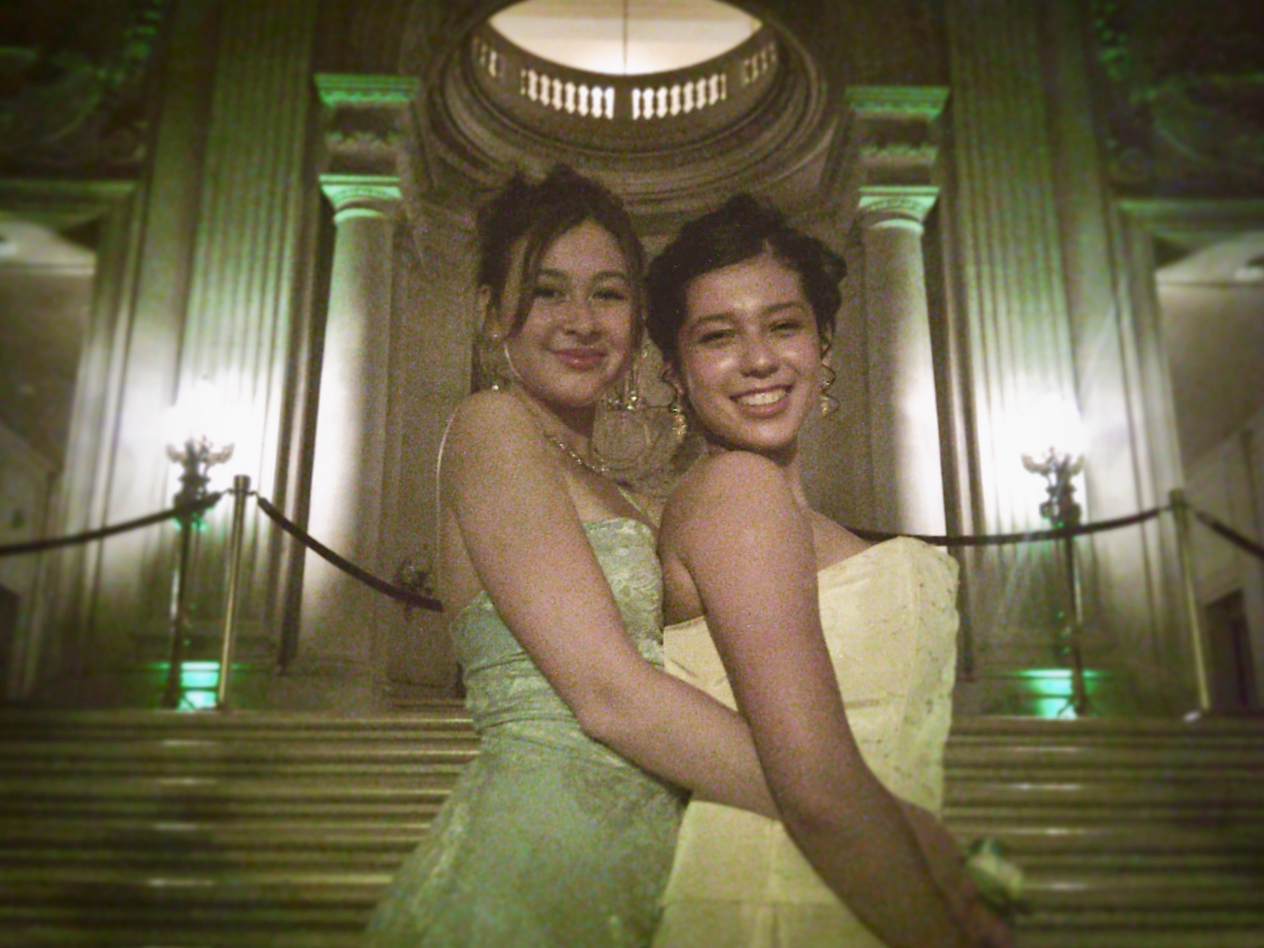 Two women in formal dresses pose in front of an elegant, large-columned building with green lighting. They are smiling and embracing each other warmly.