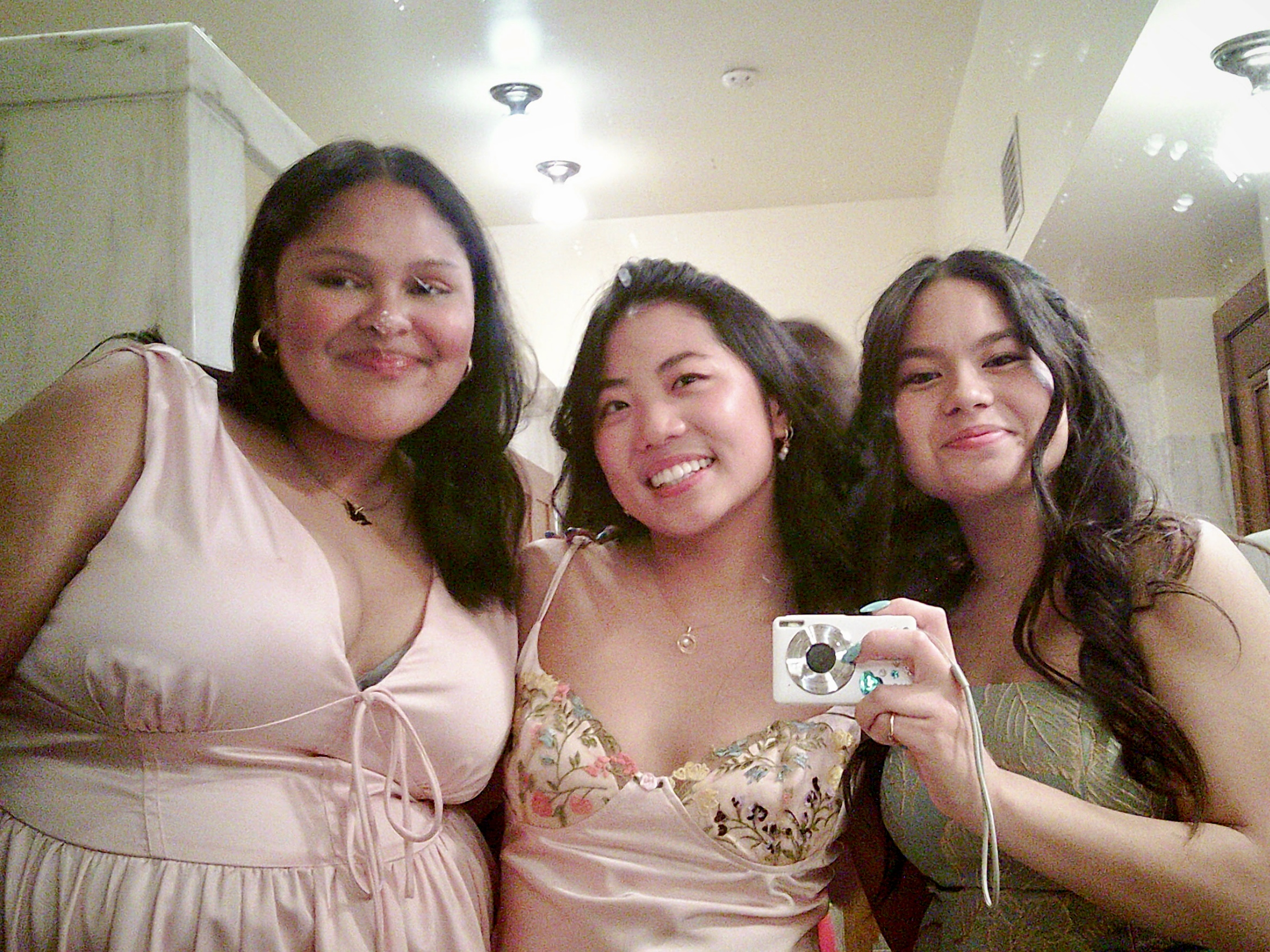 Three smiling people are taking a mirror selfie, all wearing elegant dresses. One of them holds a digital camera, and they are standing in a well-lit room.